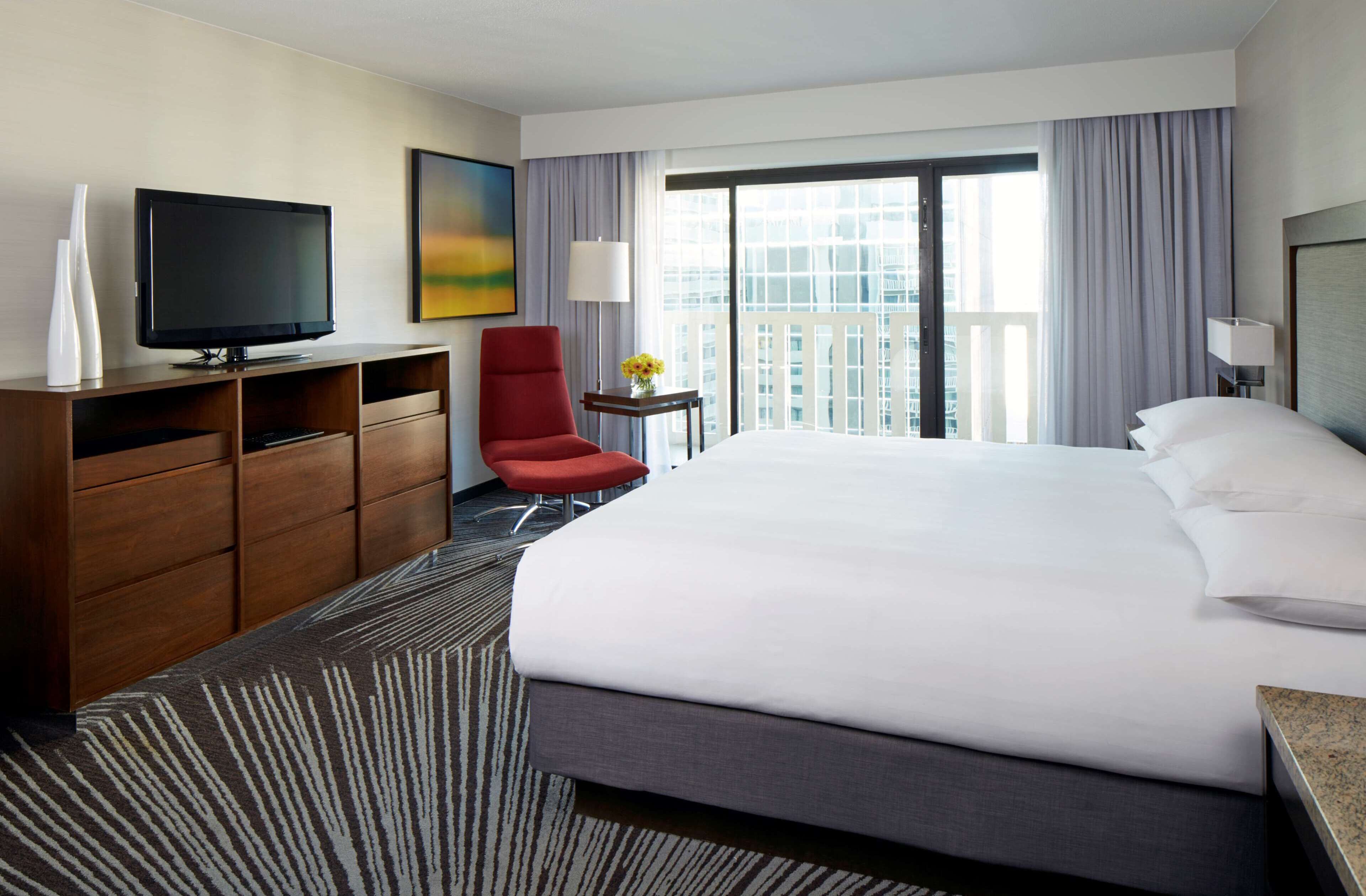 Habitación de hotel con una cama grande, televisor, una silla roja, una mesita con un jarrón y vistas a la ciudad a través de amplios ventanales.