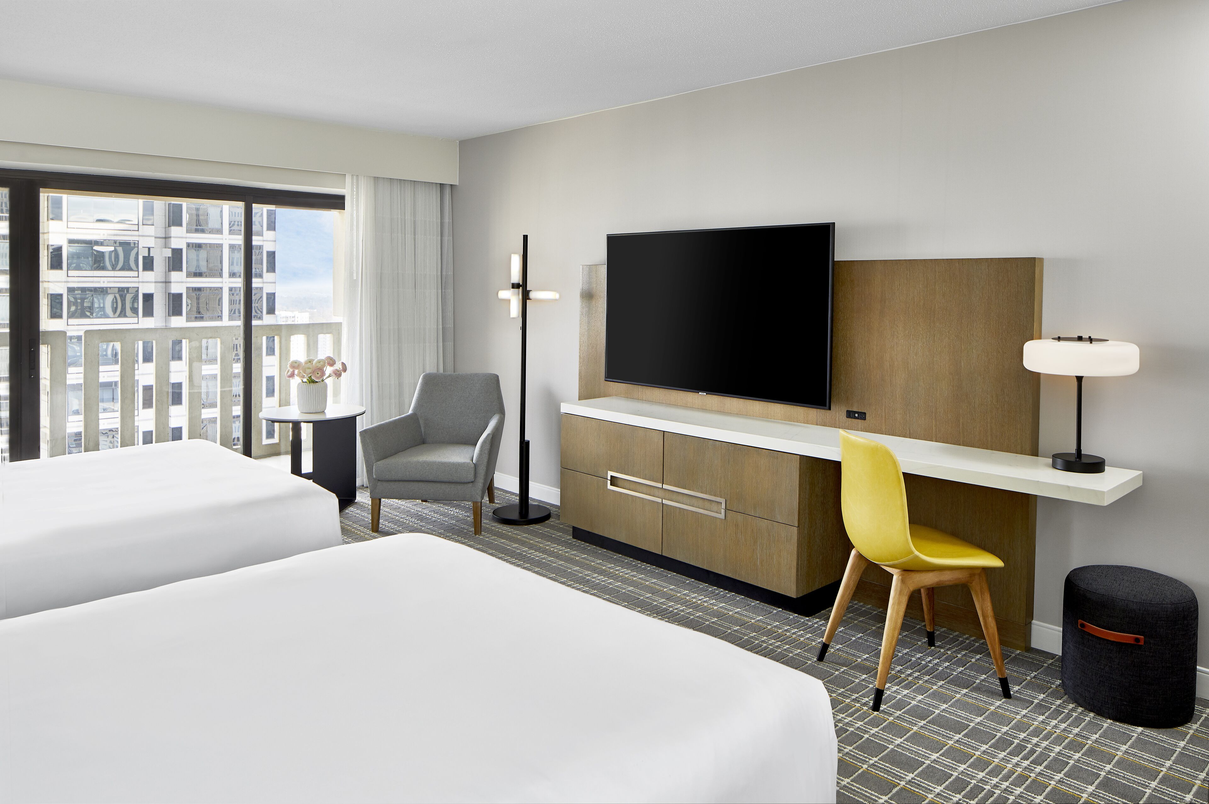 A hotel room with two beds, a desk with a flat-screen TV, a chair, a lamp, and a view of a cityscape through large windows.