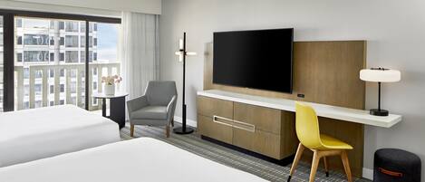 A hotel room with two beds, a desk with a flat-screen TV, a chair, a lamp, and a view of a cityscape through large windows.