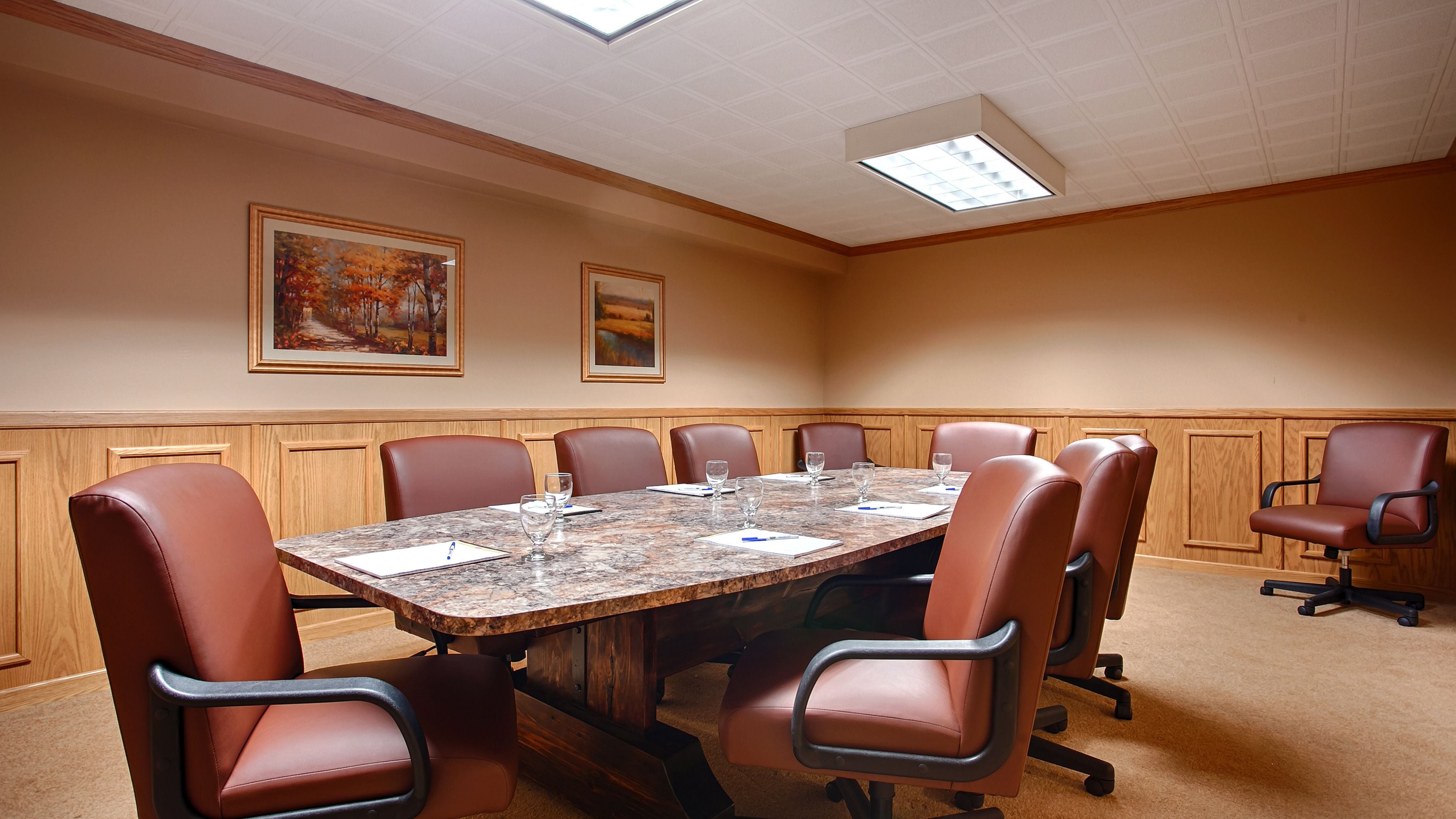 A conference room with a large table, eight leather chairs, and two framed paintings on the walls.