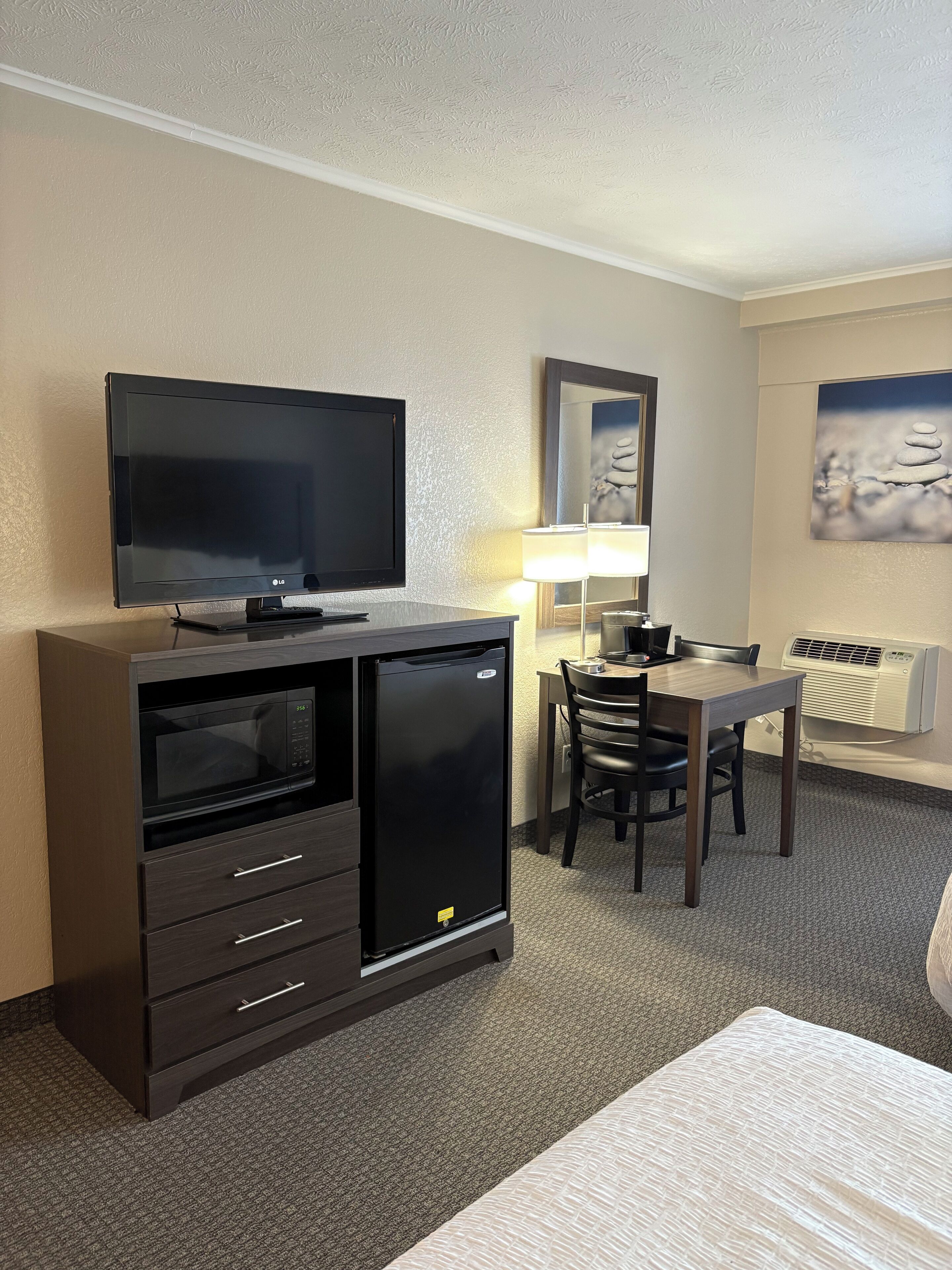 A hotel room with a flat-screen TV, a mini-fridge, a microwave, a desk with a chair, a lamp, and a painting on the wall.