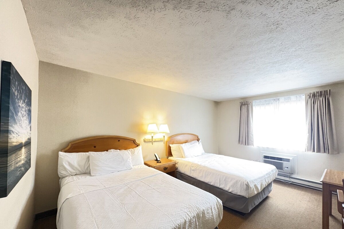 A hotel room with two beds, a nightstand with lamps, a window with curtains, and a wall-mounted air conditioner.