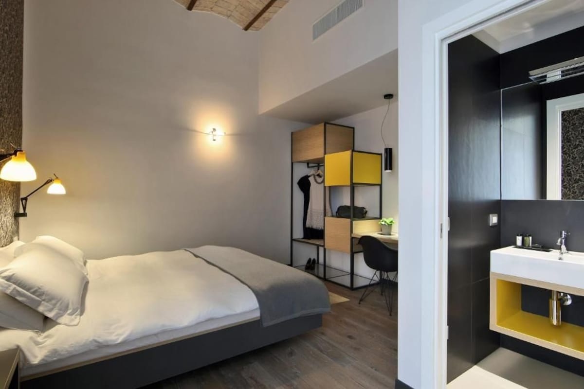 A modern bedroom with a bed, bedside lamps, a desk, and a bathroom with a mirror and sink.