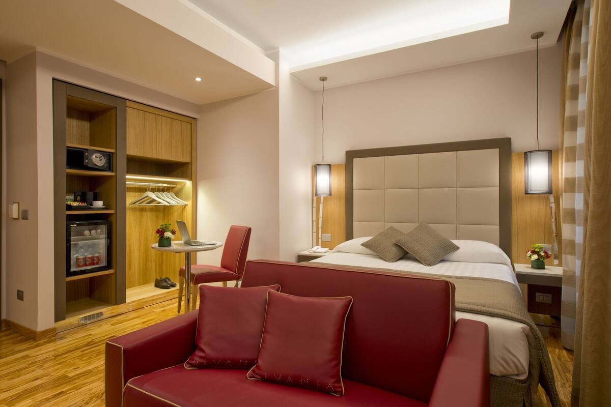 A hotel room with a bed, a red sofa, a wardrobe, a desk with a chair, and a small table with a vase.