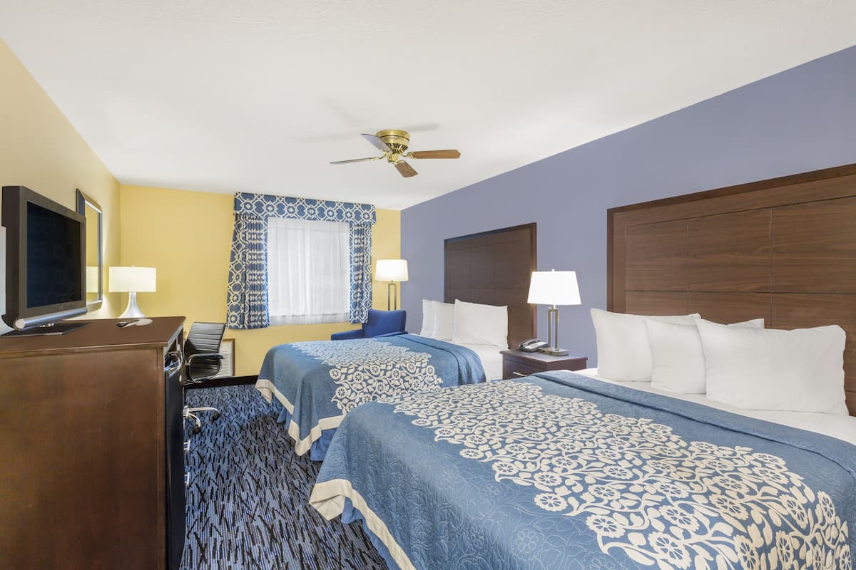 Quarto de hotel com duas camas, uma escrivaninha com televisão, ventoinha no teto e janela com cortinas estampadas.