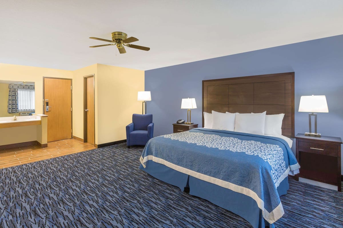 Quarto de hotel com uma cama grande, duas mesas de cabeceira com luminárias, uma cadeira, uma escrivaninha com espelho e um ventilador de teto.