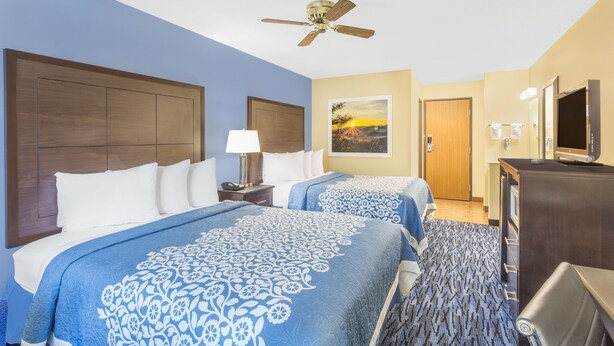 A hotel room with two beds, a ceiling fan, a painting, a TV, and a desk.