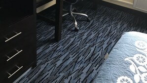 A hotel room with a desk, chair, and a bed. There is a lamp on the desk, a clock, and a mirror on the wall.