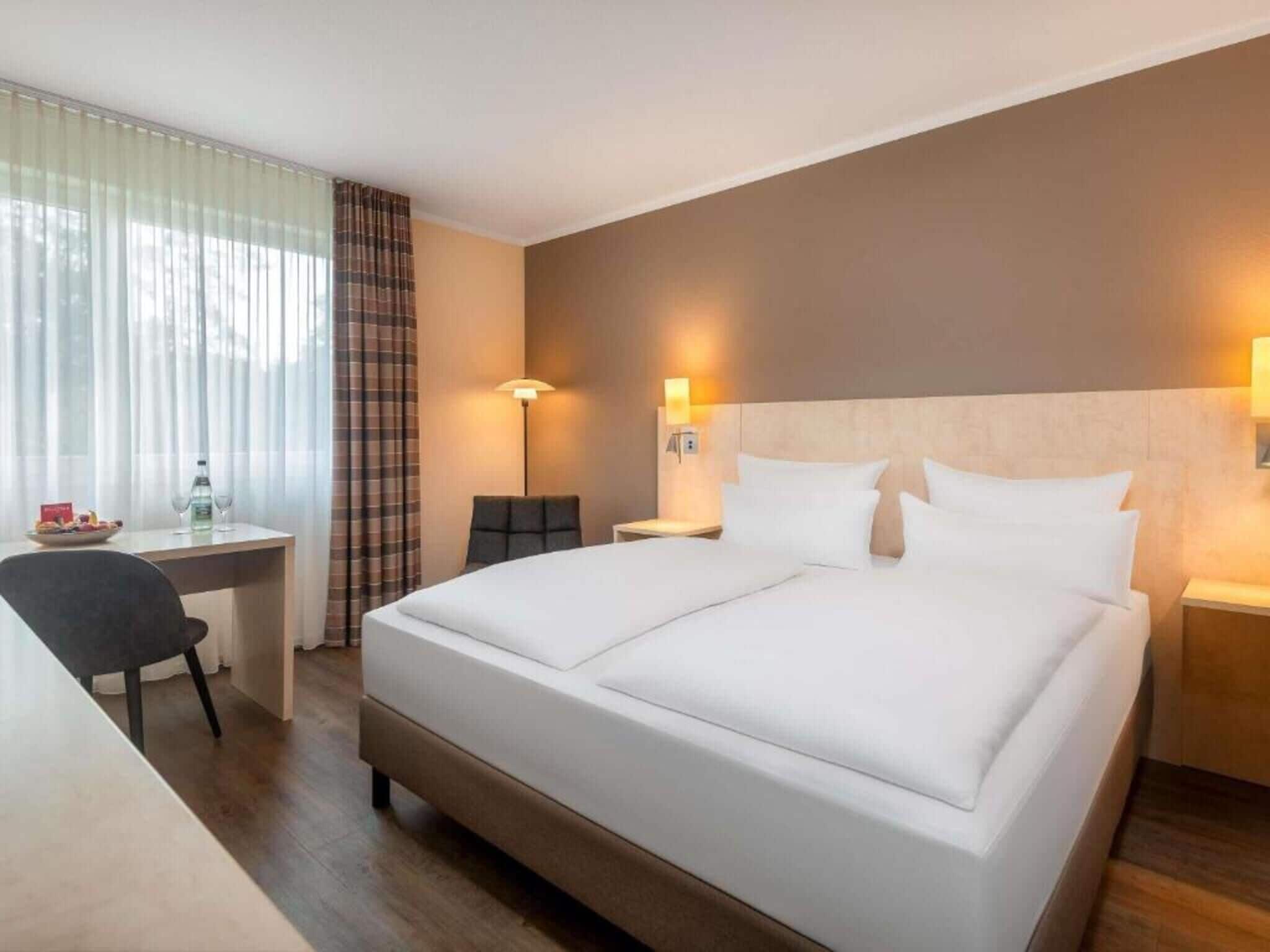 A hotel room with a double bed, a desk with a chair, a lamp, and a window with curtains.