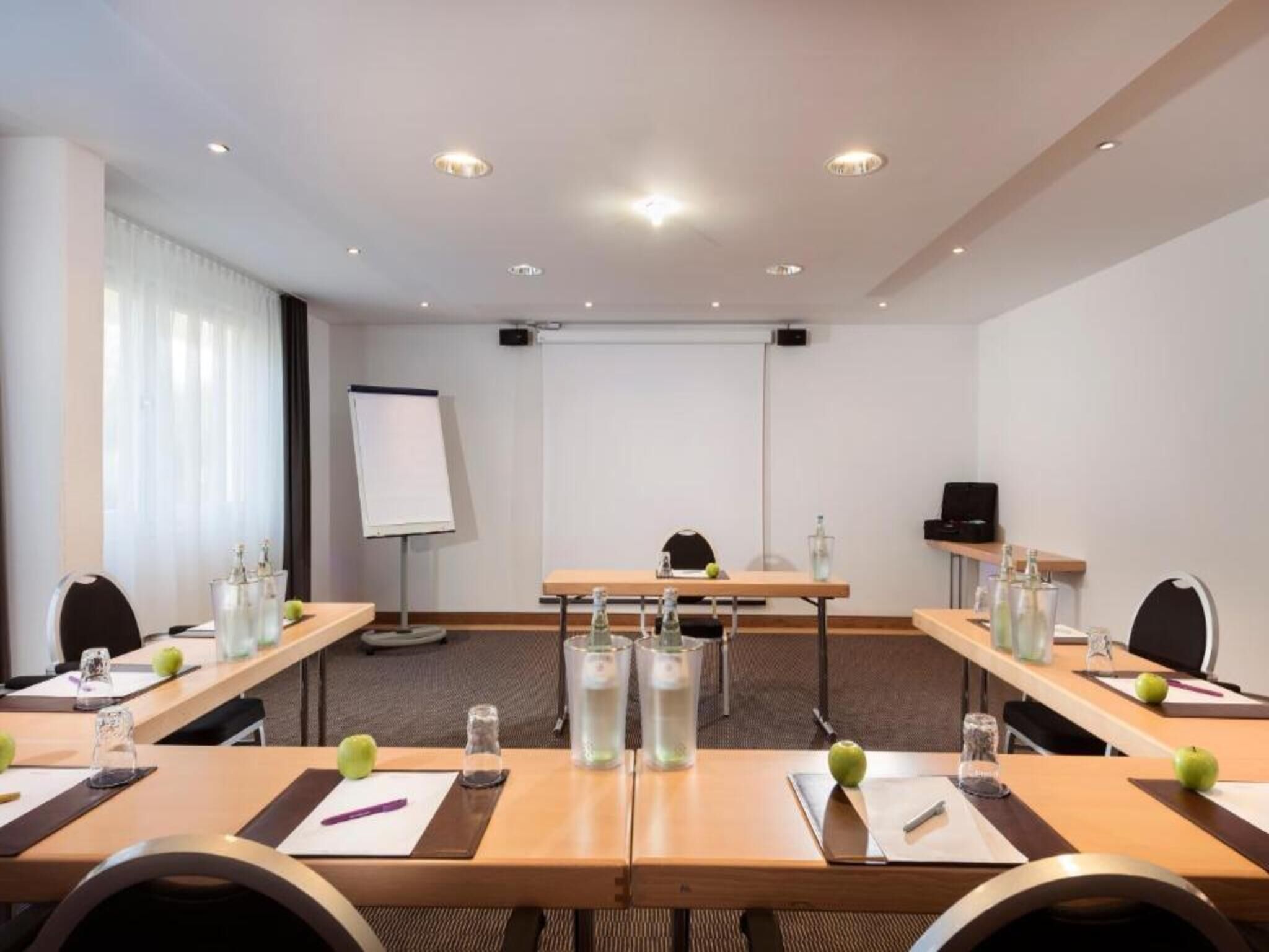 Una sala de conferencias con mesas, sillas, una pantalla de proyector y una pizarra.