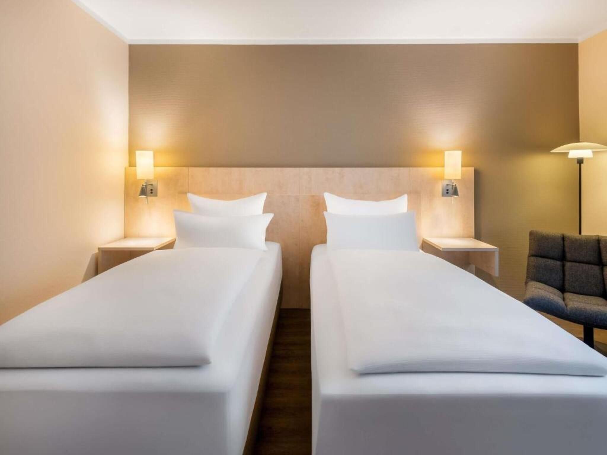 A hotel room with two single beds, nightstands, wall-mounted lamps, and a sofa.