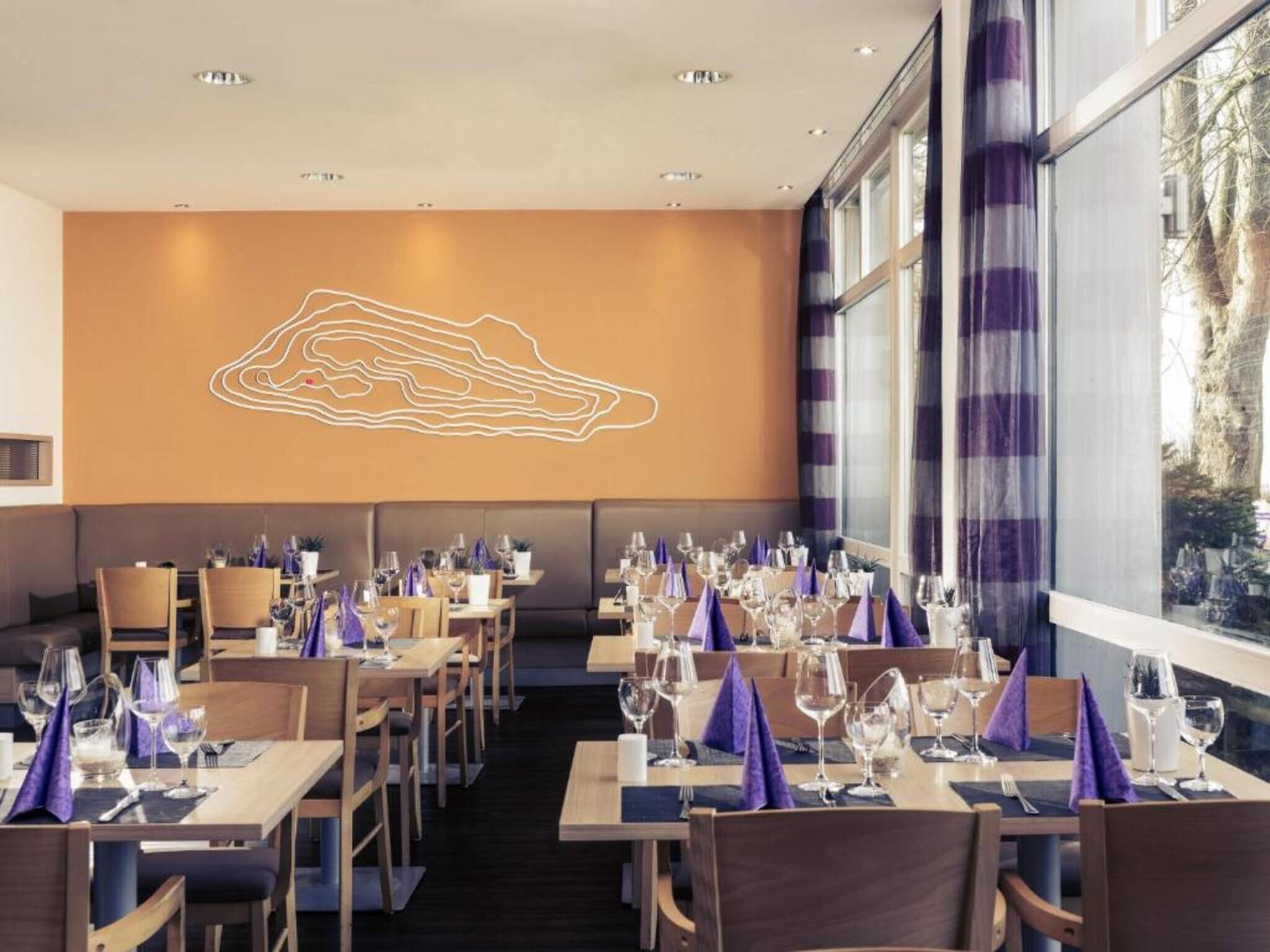 A restaurant with tables set for dining, a large window with curtains, and a wall with a geometric design.