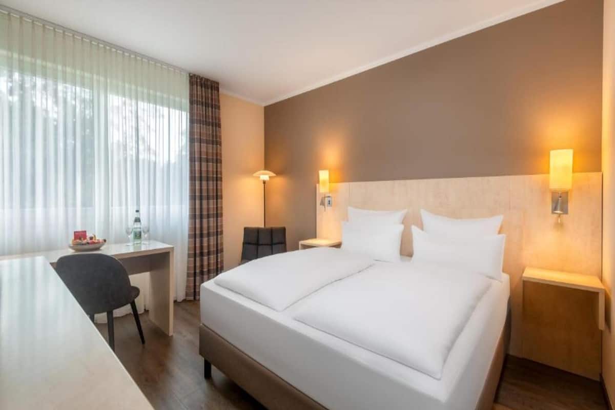 Una habitación de hotel con cama, mesitas de noche, escritorio, silla y ventana con cortinas.