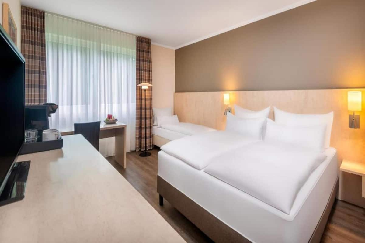 Una habitación de hotel con cama, mesitas de noche, un escritorio con silla, televisión y ventana con cortinas.