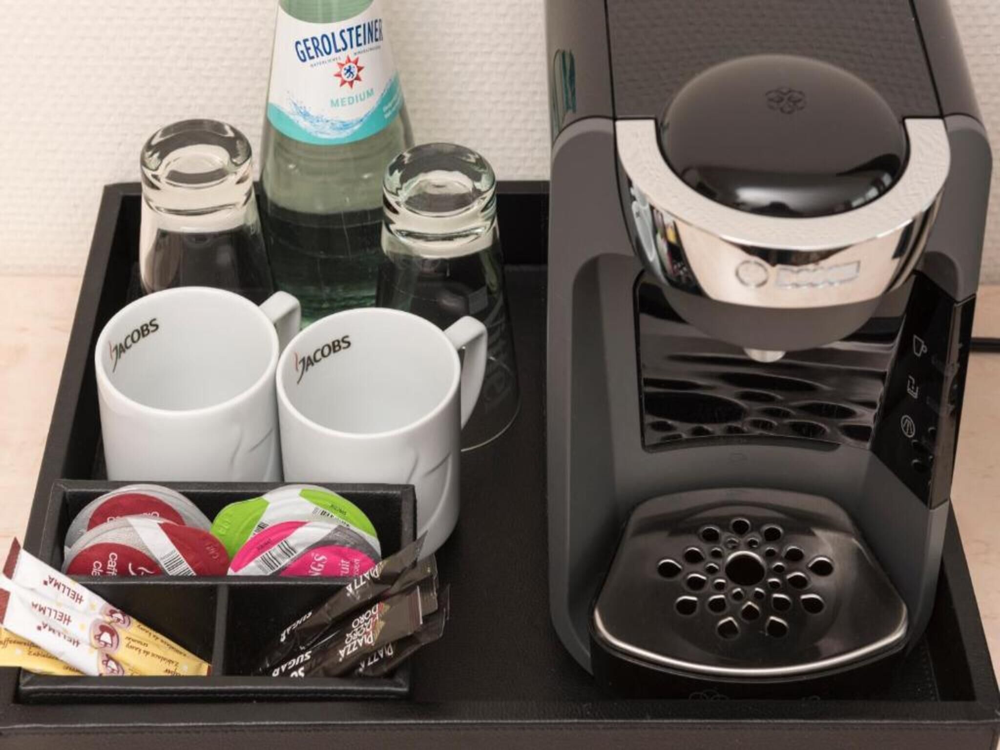 A coffee machine, two coffee mugs, two glass bottles, and a tray with coffee pods and a packet of sugar.