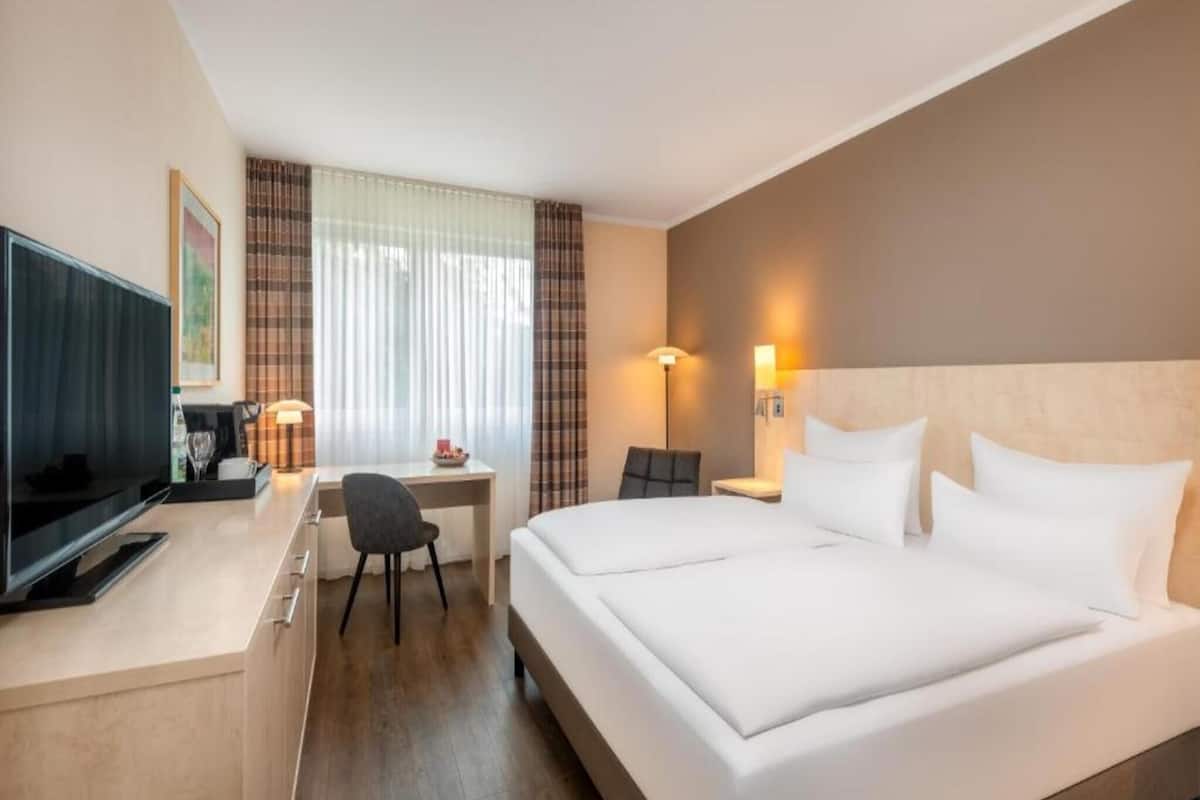 Habitación de hotel con cama, escritorio, silla, televisor y cortinas.