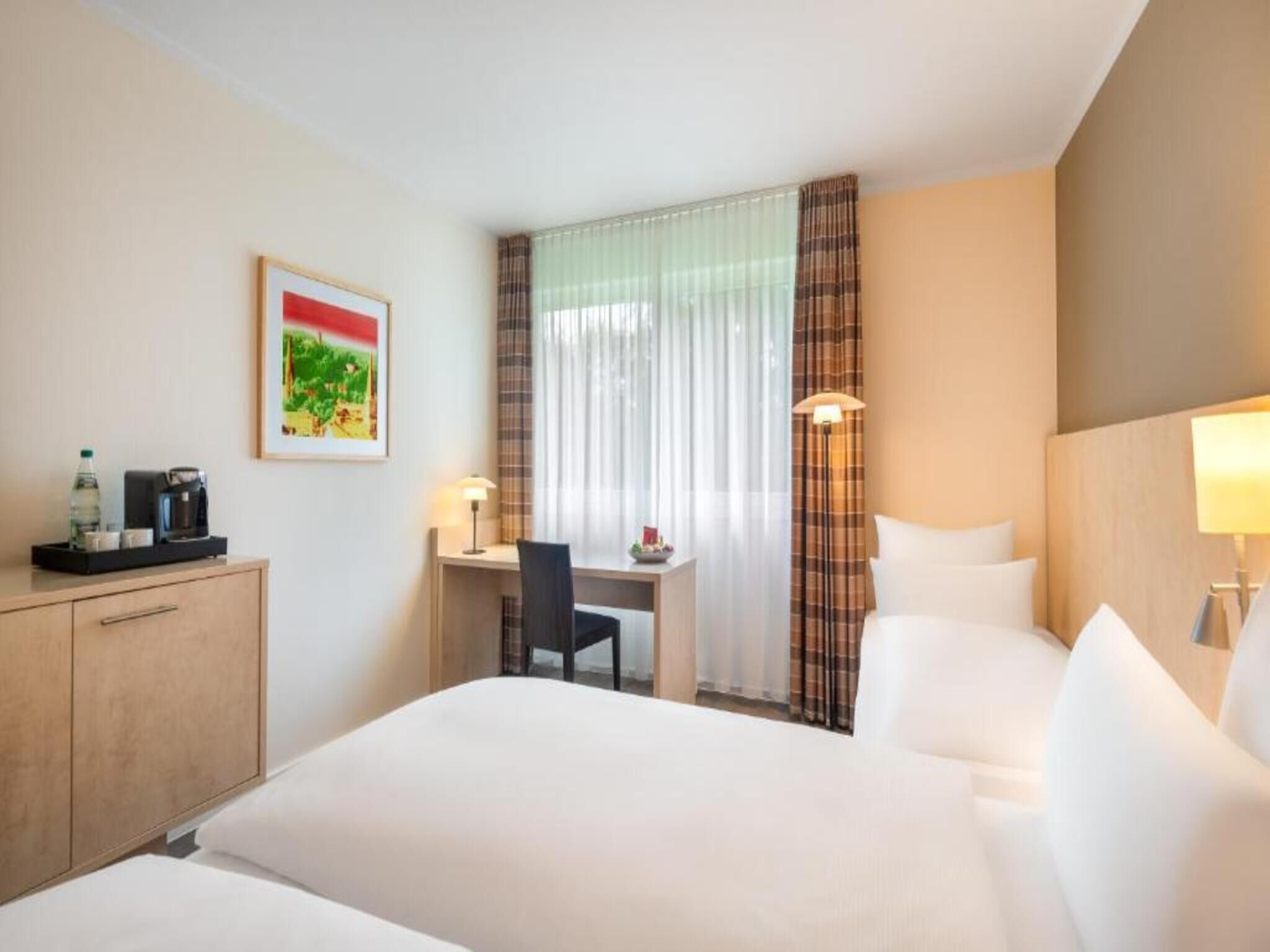 A hotel room with a bed, a desk, a chair, a coffee machine, and a painting on the wall.