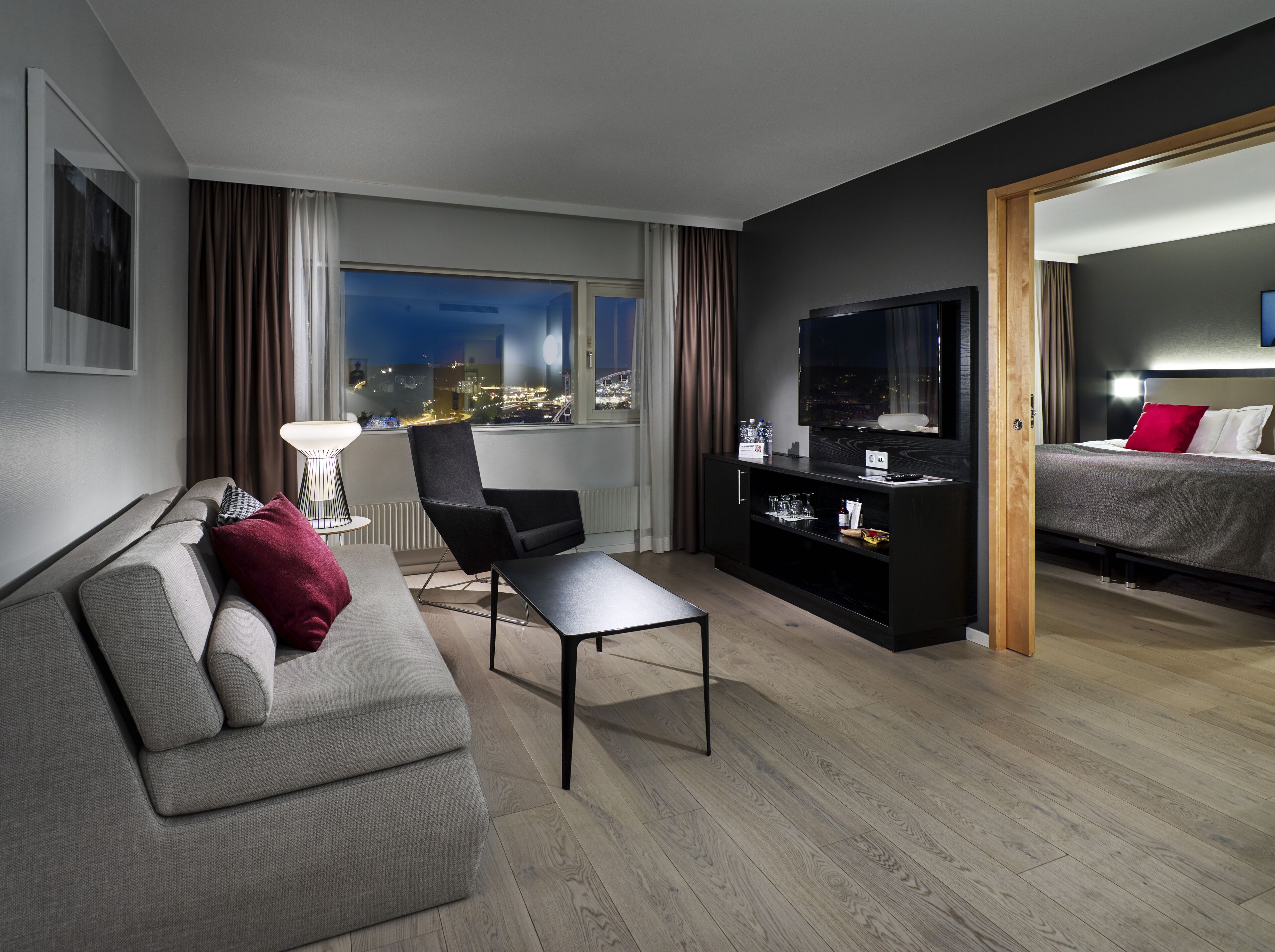 A modern hotel room with a sofa, armchair, and a bed. There is a TV on a stand and a city view through the window.