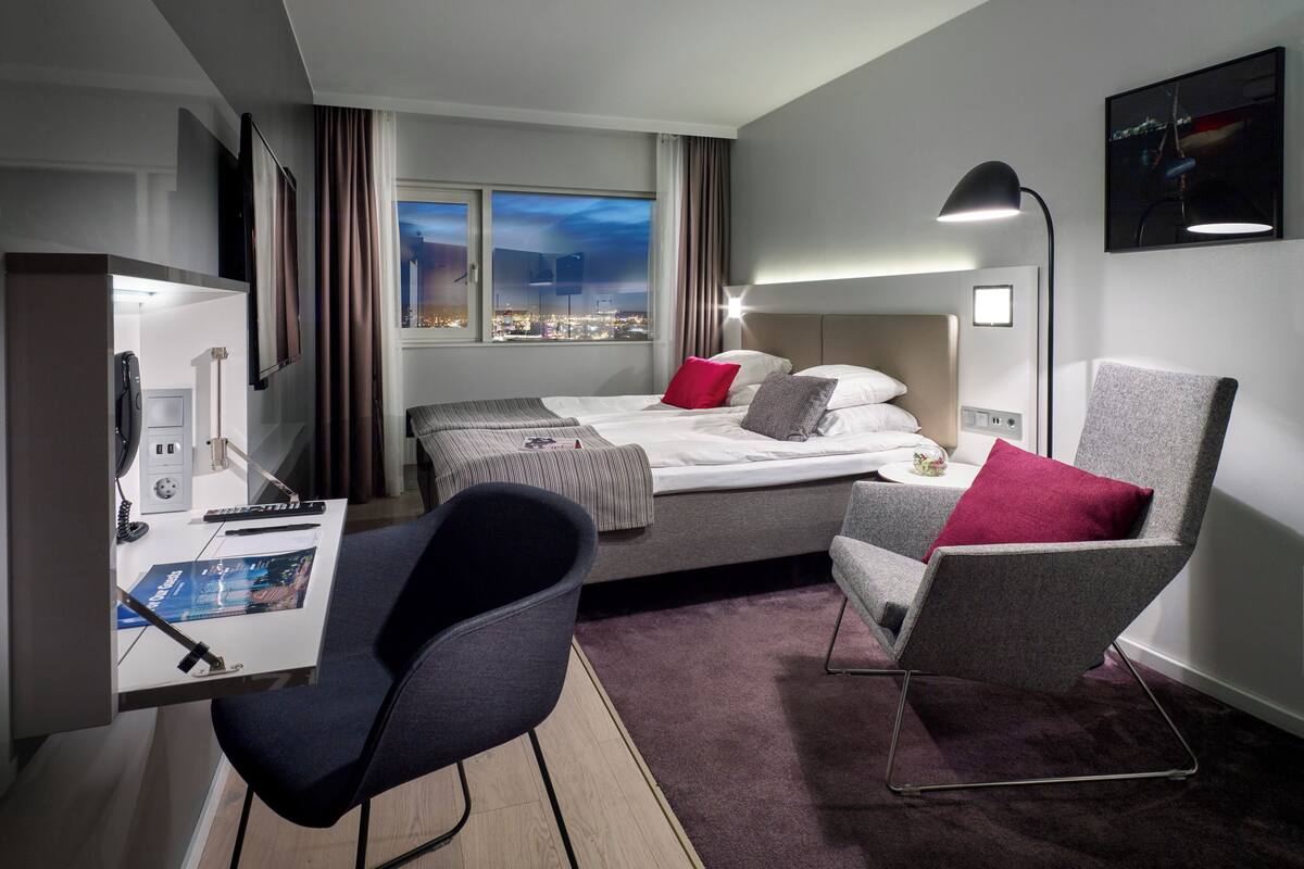 Ein Hotelzimmer mit Bett, Schreibtisch, Stuhl und Blick auf die Stadt durch das Fenster.