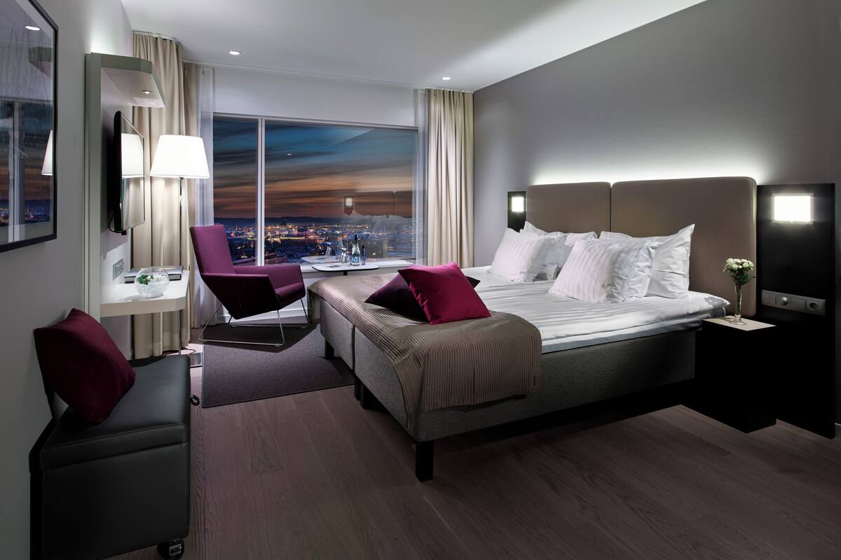 Ein modernes Hotelzimmer mit einem großen Bett, einem Schreibtisch, einem Sessel und Blick auf eine Stadtlandschaft in der Abenddämmerung.