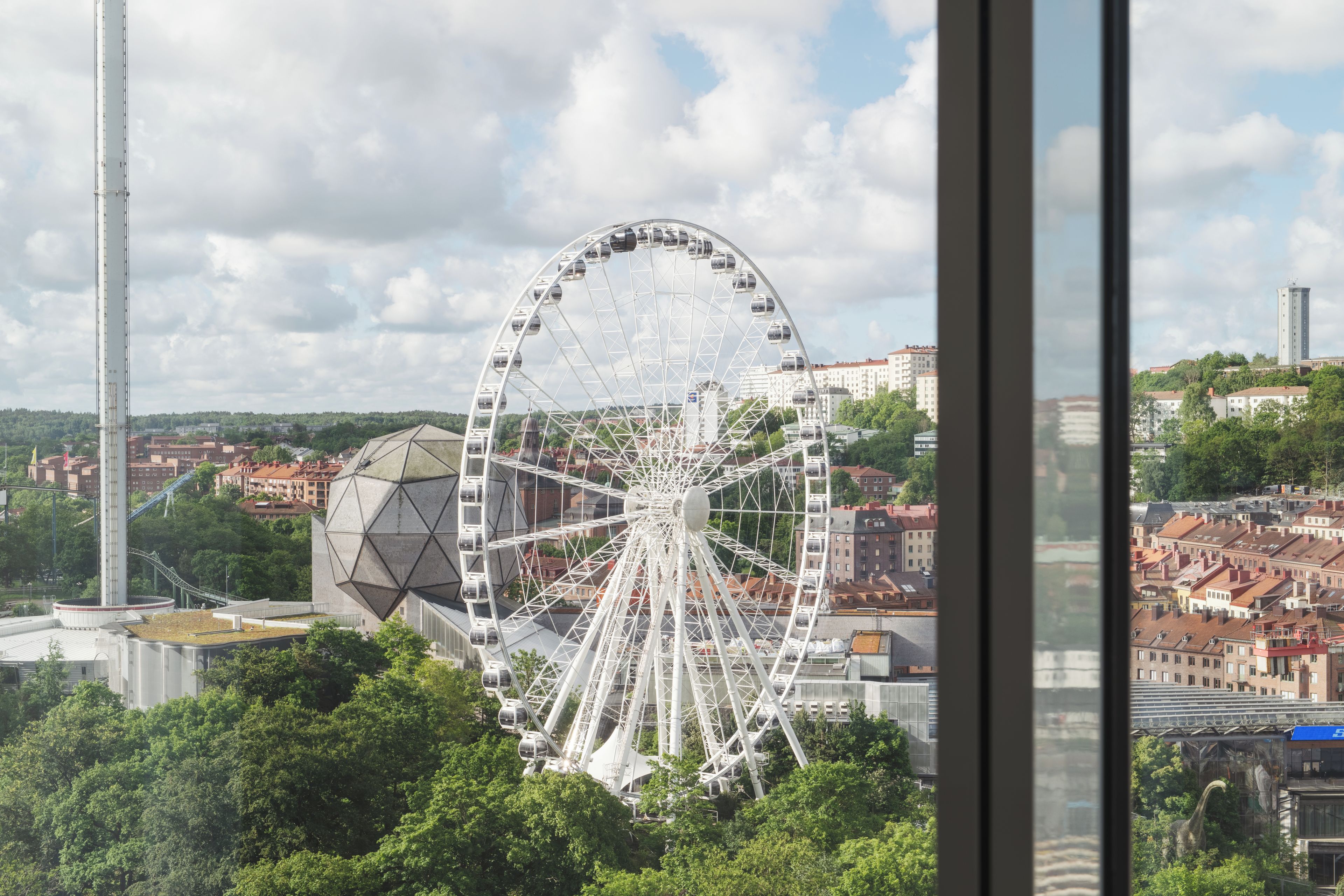 A Ferris wheel with a transparent cabin structure, surrounded by urban buildings and greenery.