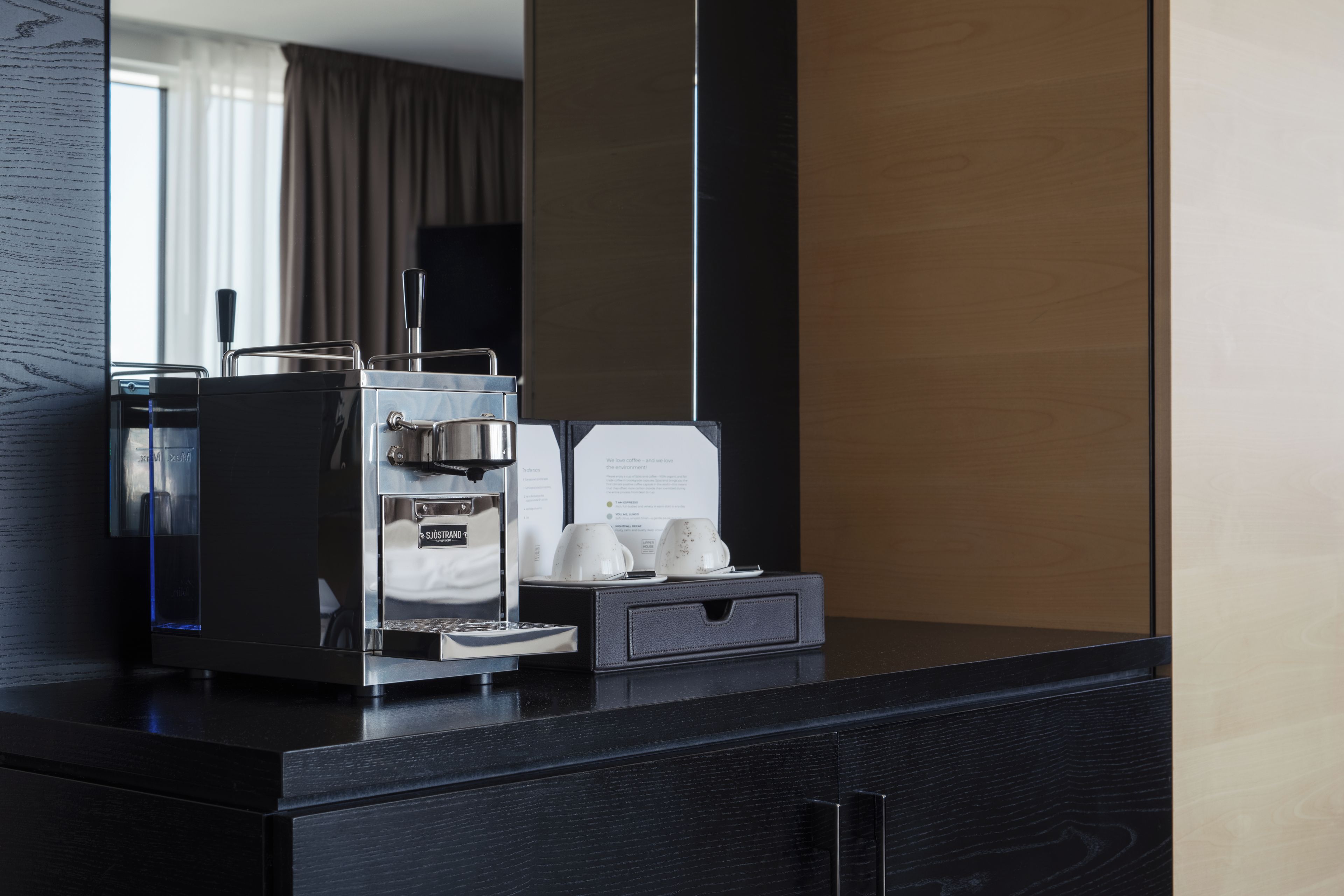 A hotel minibar with a coffee machine, a glass of water, and a menu.