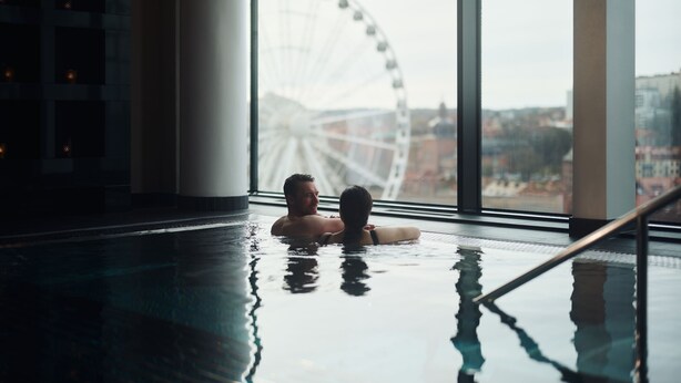 Un homme et une femme se détendent dans une piscine, et on peut voir une grande grande roue à travers la fenêtre.