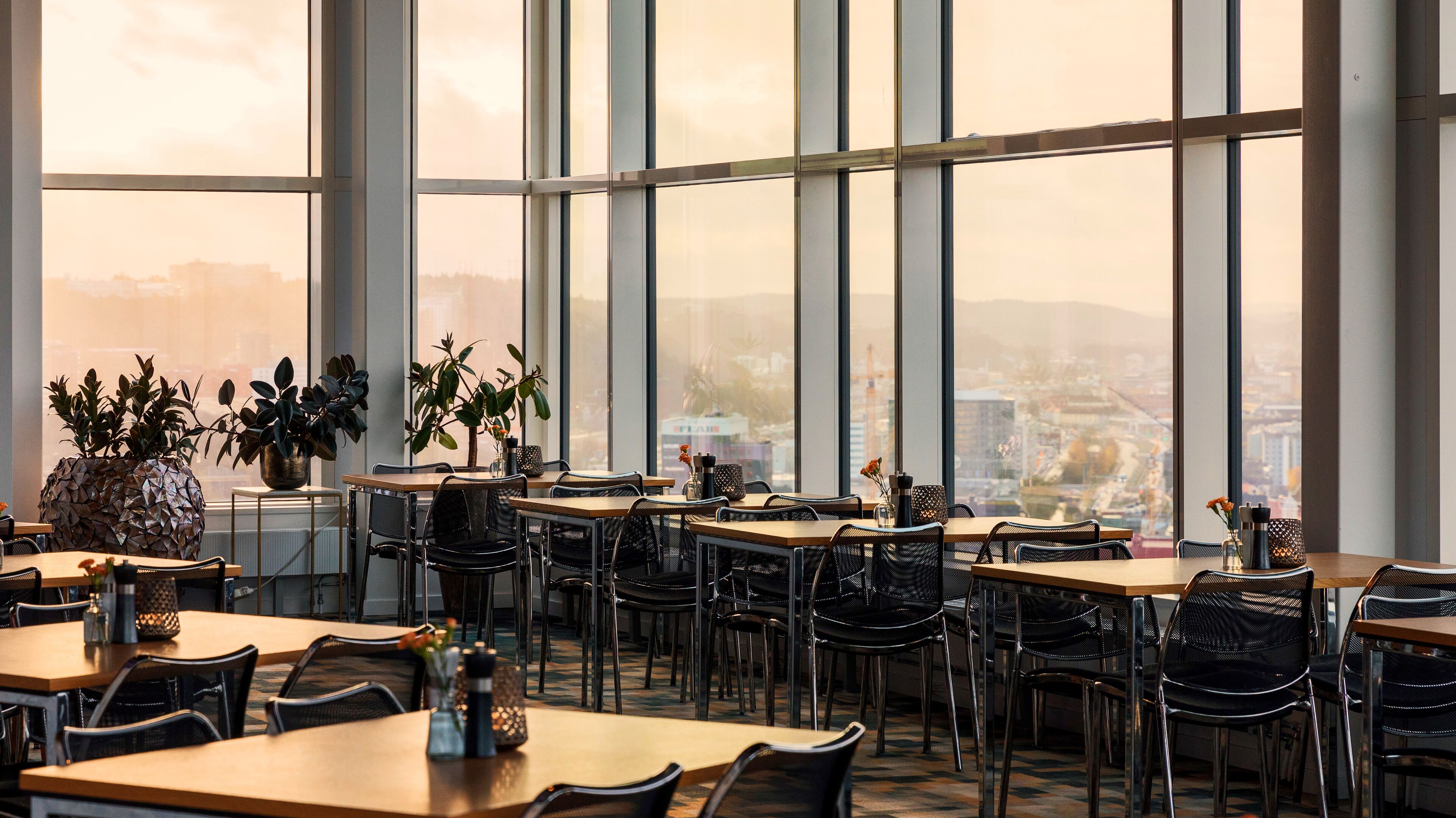 Een modern restaurant met grote ramen, meerdere tafels en stoelen, met uitzicht op de stad.