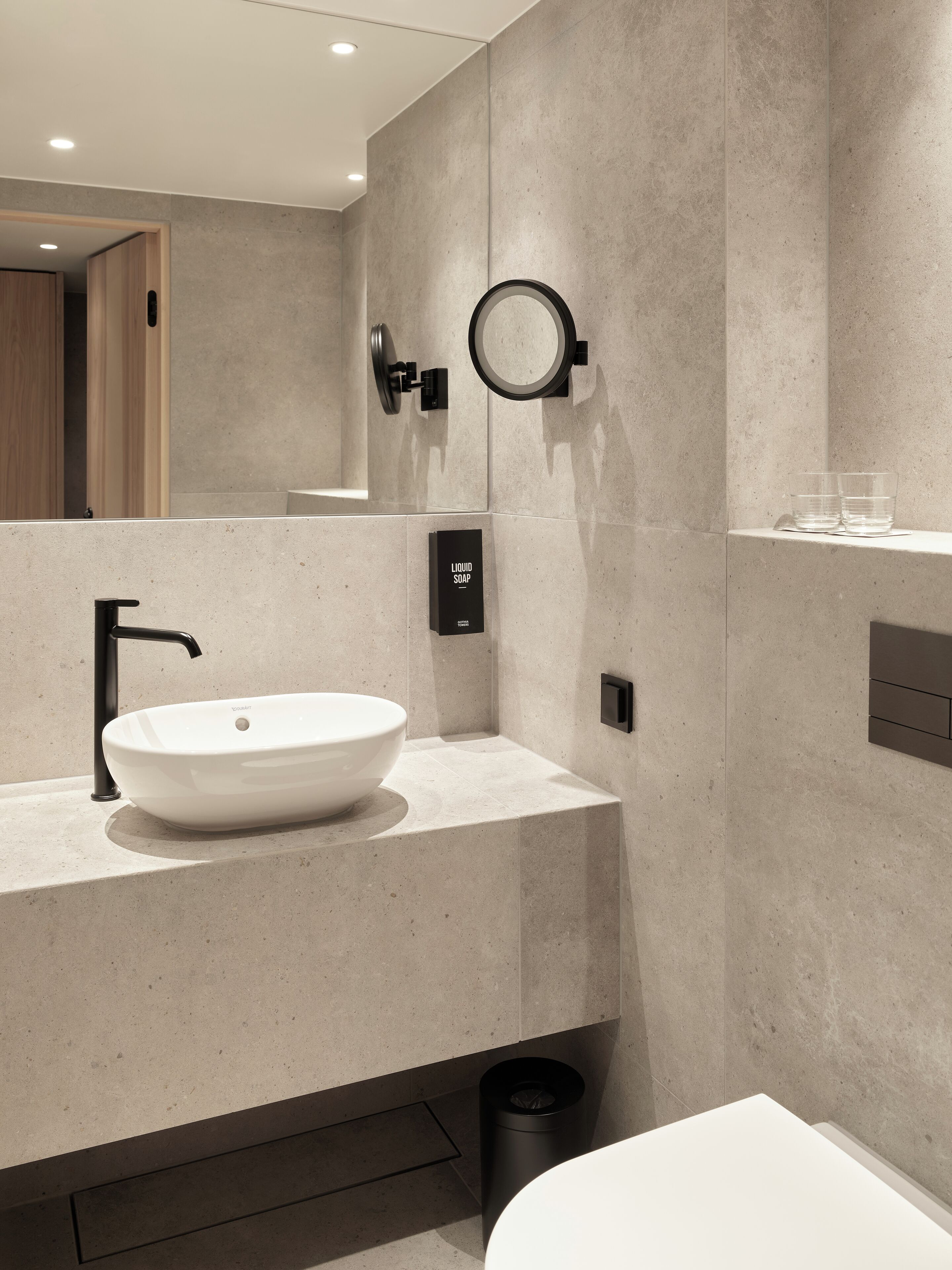 Un bagno moderno con un lavandino bianco capiente, uno specchio rotondo e un cestino nero.