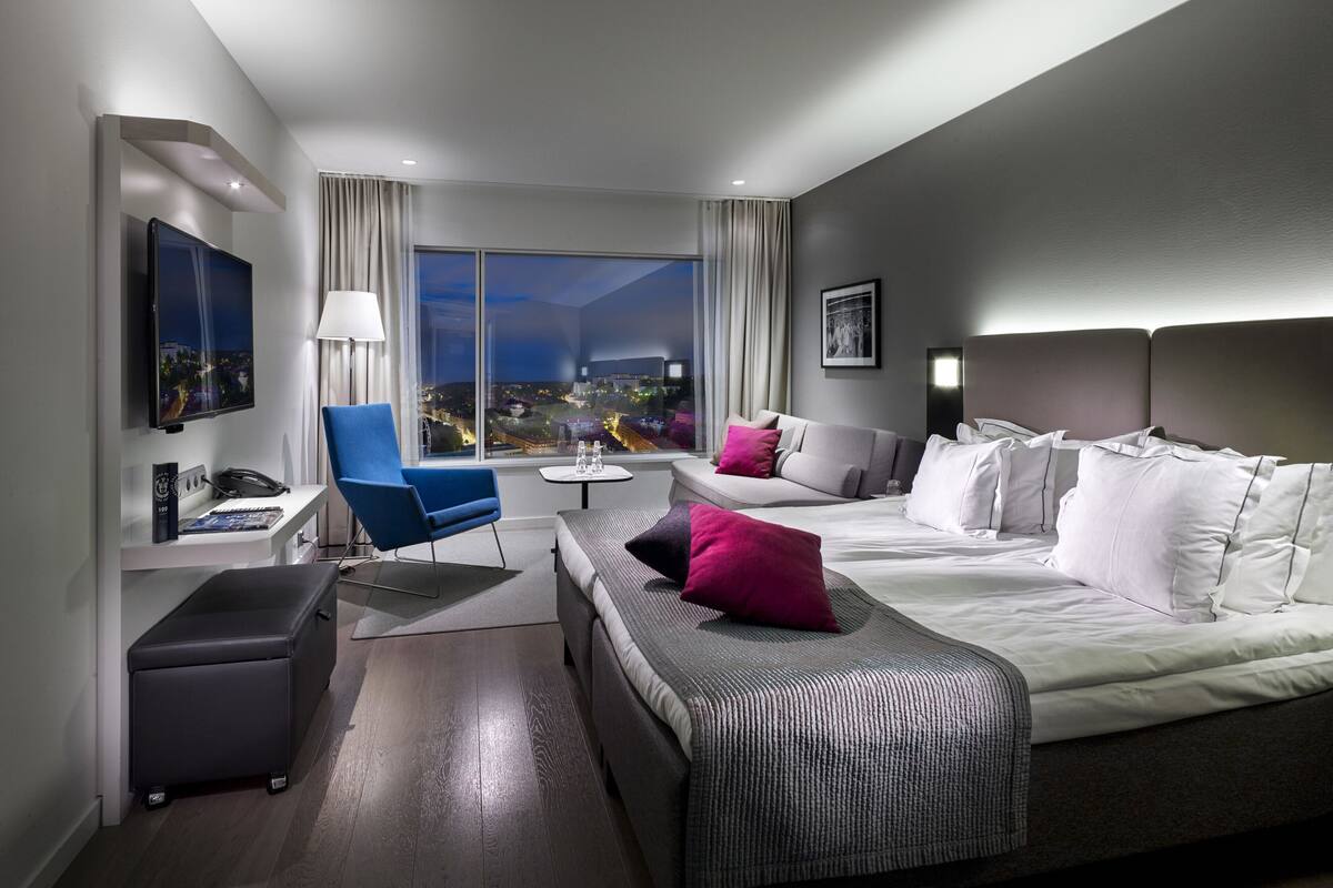 Ein modernes Hotelzimmer mit einem großen Bett, einer Couch, einem Schreibtisch mit Computer und einem Sessel. Ein Fenster bietet Blick auf die Stadt.