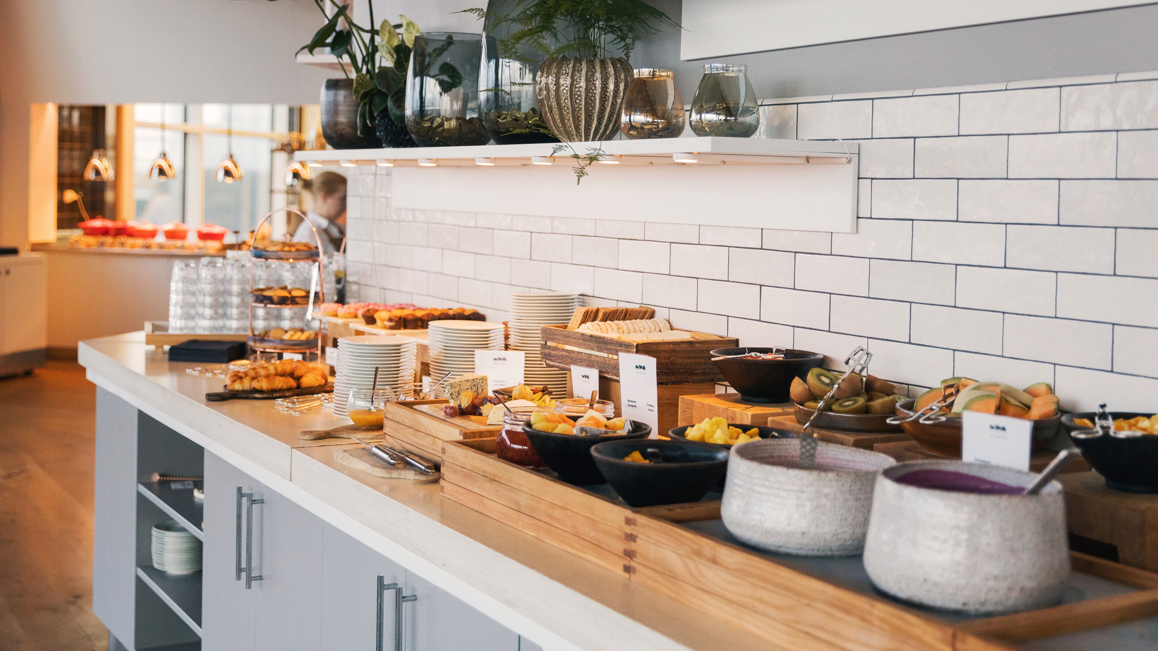 A buffet with various food items, including fruits, cheeses, and pastries, arranged on a counter with a tiled backsplash and a plant shelf above.