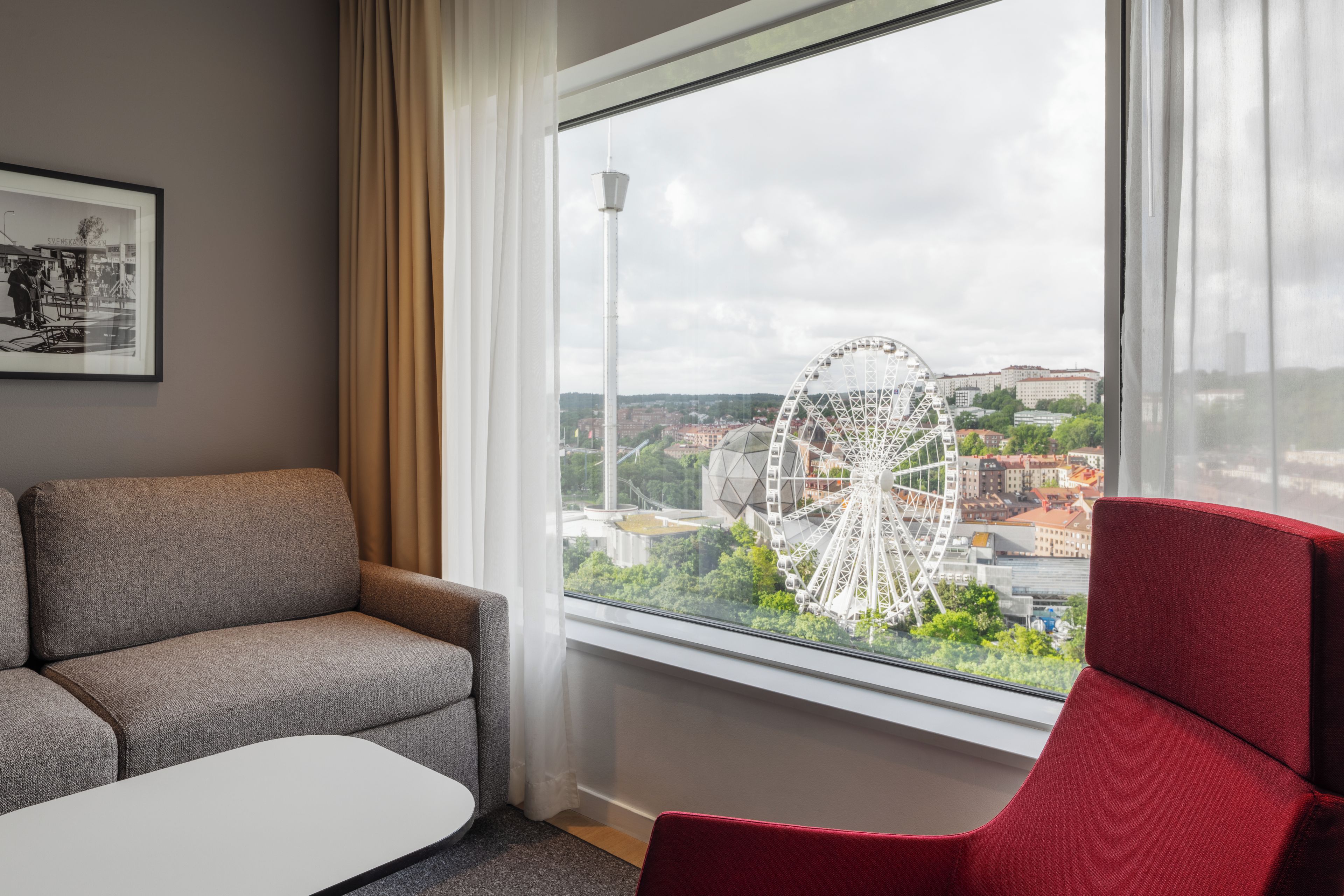A room with a sofa, a red chair, a small table, and a window with a view of a Ferris wheel and buildings.