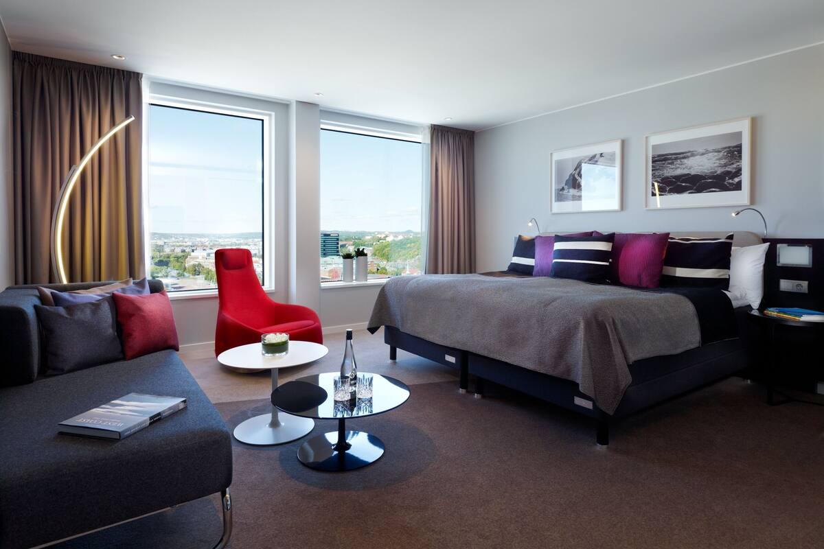 Ein modernes Hotelzimmer mit einem Bett, einem roten Sessel, einer Couch und einem kleinen Tisch mit einem Buch und Gläsern.