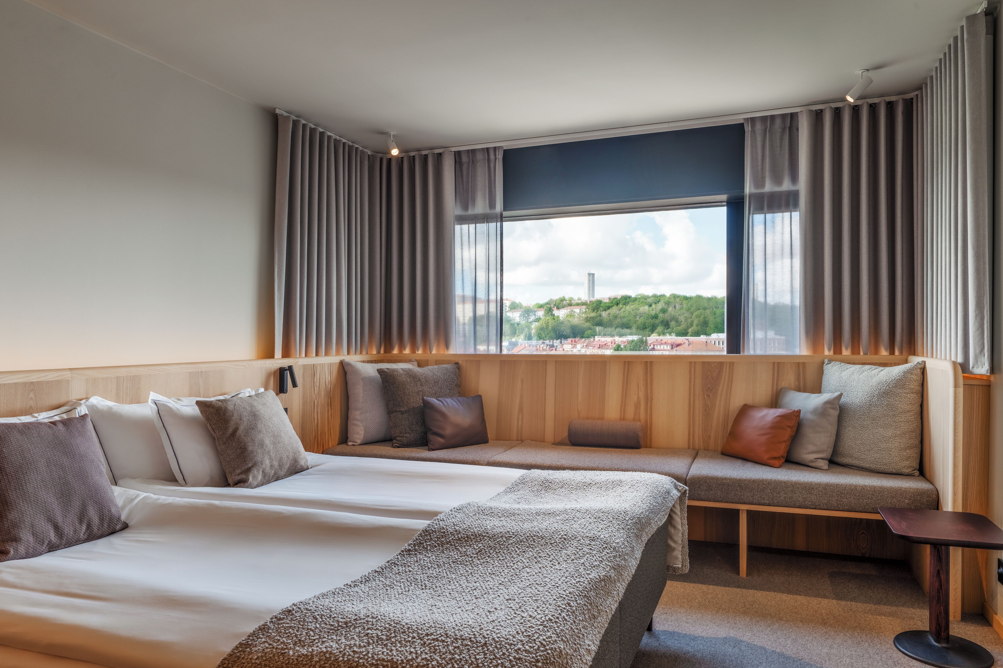 Une chambre d’hôtel avec un grand lit, un coin salon avec des coussins, une petite table et une fenêtre avec des rideaux.
