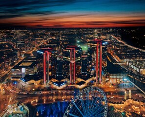 A nighttime cityscape with prominent skyscrapers, a Ferris wheel, and a large building with a distinctive red-lit structure.