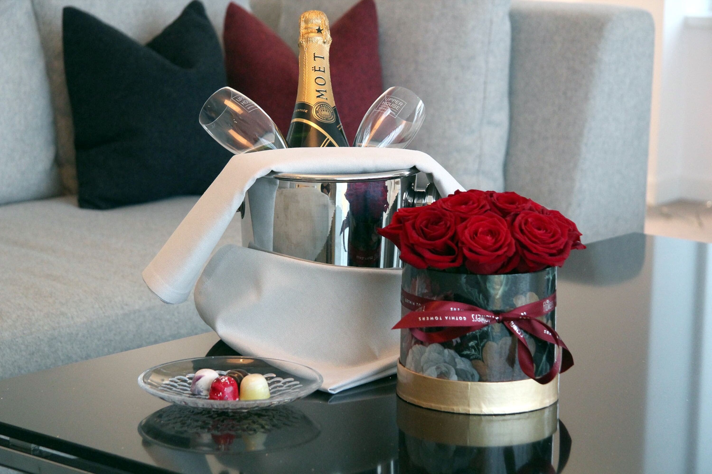 A champagne bottle in a bucket, a glass, and a vase with red roses on a table.