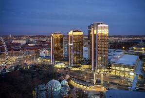 Paisagem urbana ao entardecer com arranha-céus iluminados, incluindo as Gotham Towers, e uma roda gigante em primeiro plano.