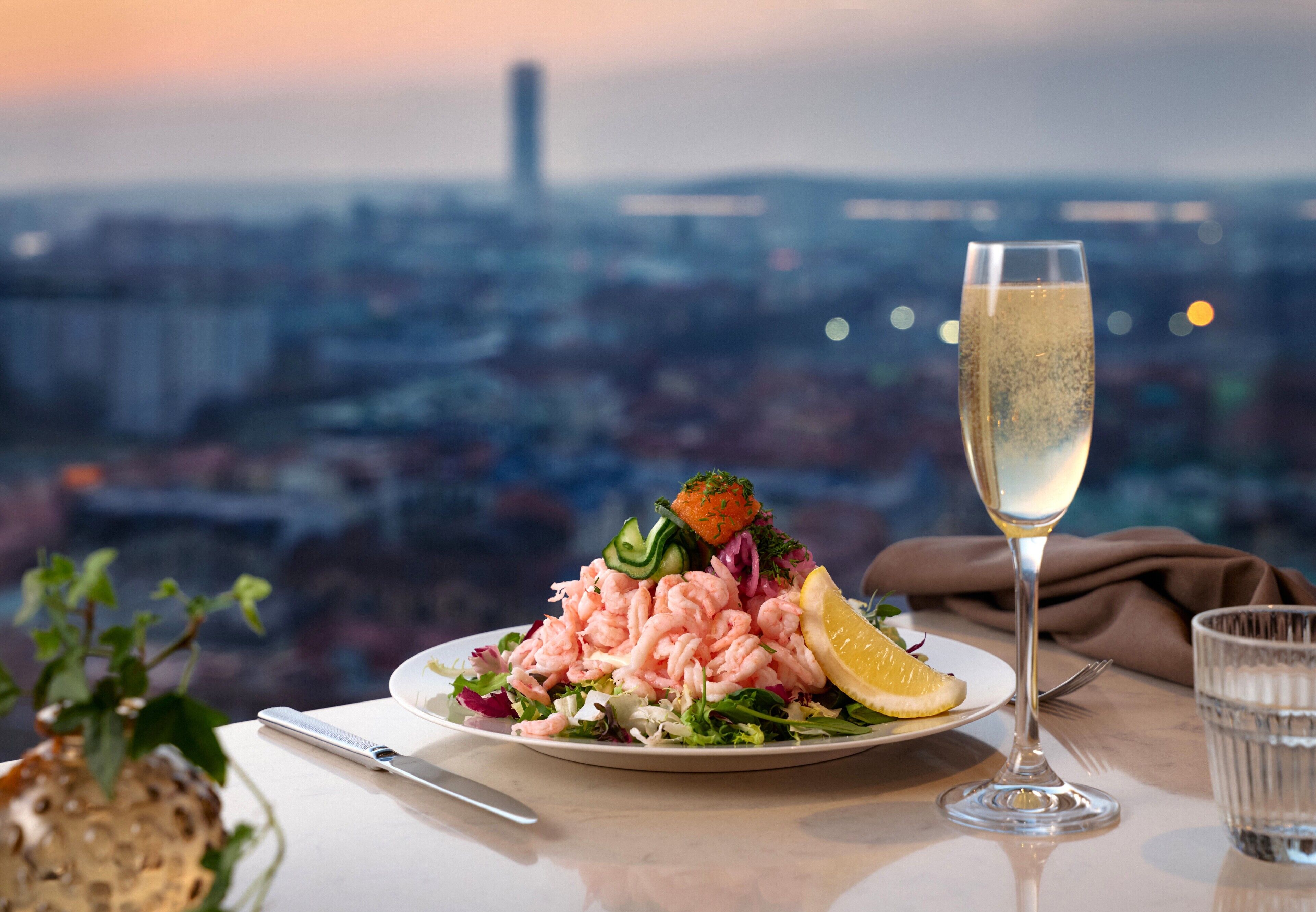 A plate of shrimp salad with a lemon wedge, a glass of champagne, and a city view in the background.