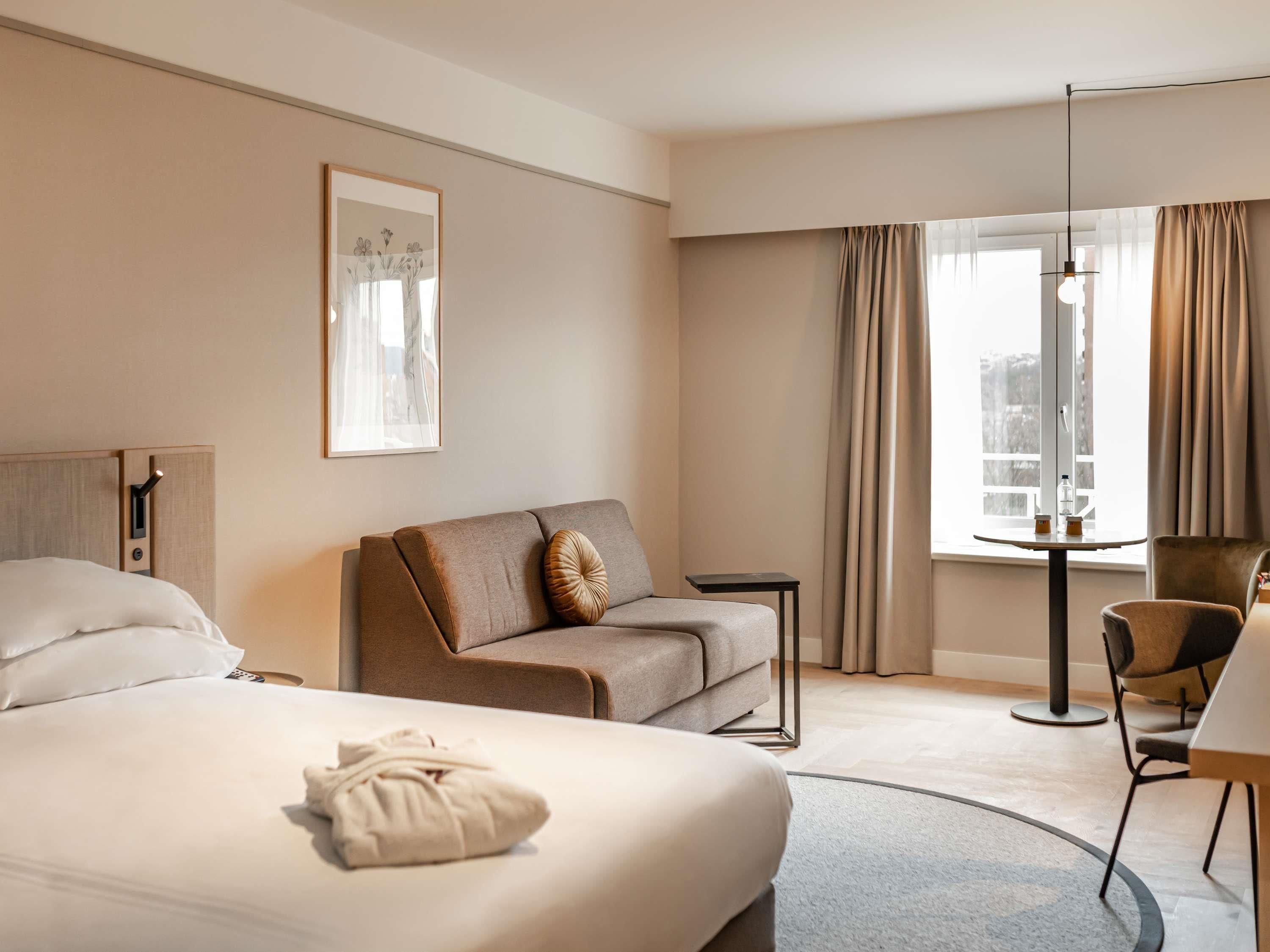 Una camera d'albergo con un letto, un divano, un tavolino, una sedia, una finestra con tende e un quadro appeso al muro.