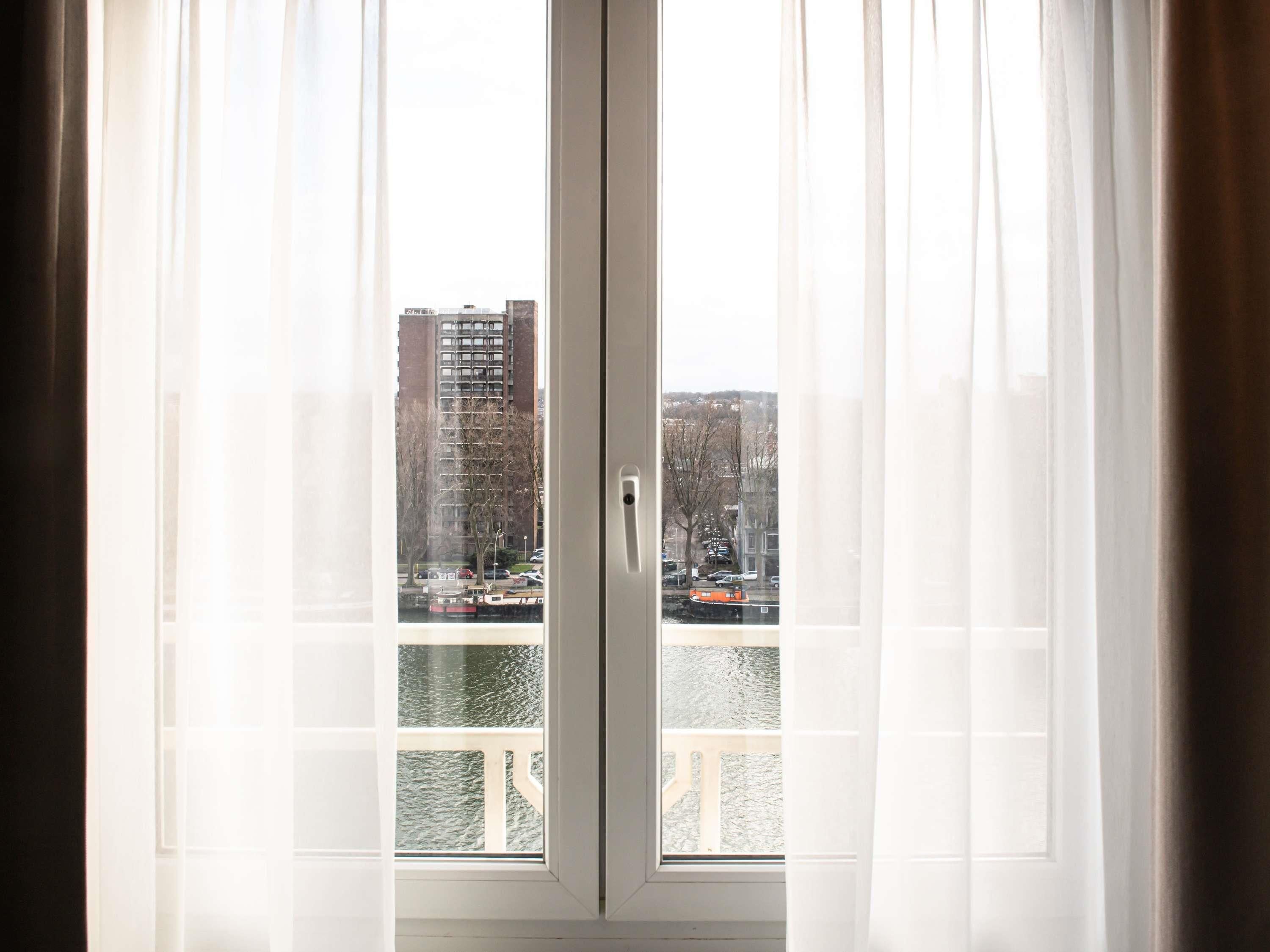 Vista a través de una ventana con cortinas translúcidas, mostrando un paisaje urbano con edificios y una masa de agua.