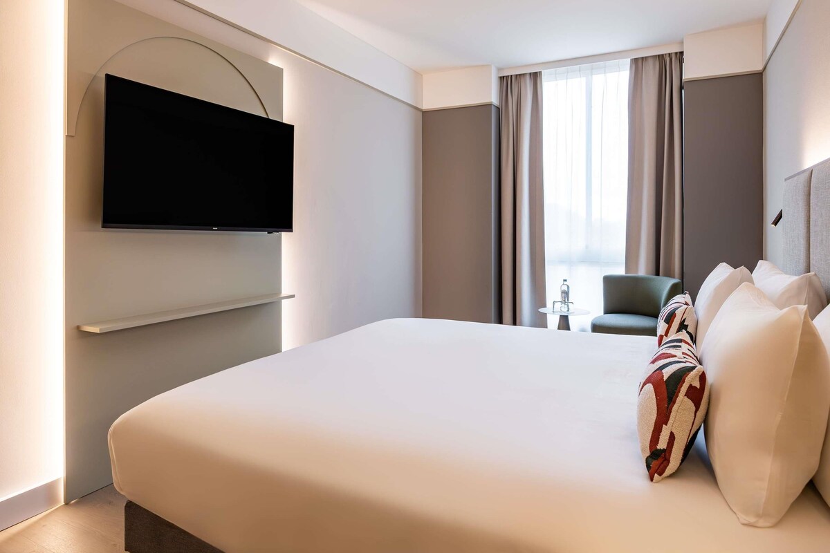 A modern hotel room with a large bed, a flat-screen TV mounted on the wall, a small table, and a chair.