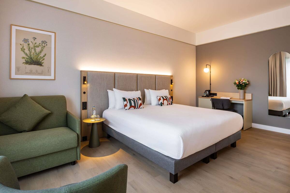 A hotel room with a bed, a green armchair, a desk with a lamp, a mirror, and a framed botanical illustration on the wall.