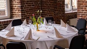 Een ronde eettafel met een wit tafelkleed, compleet met wijnglazen, bestek en een bloemenvaas.