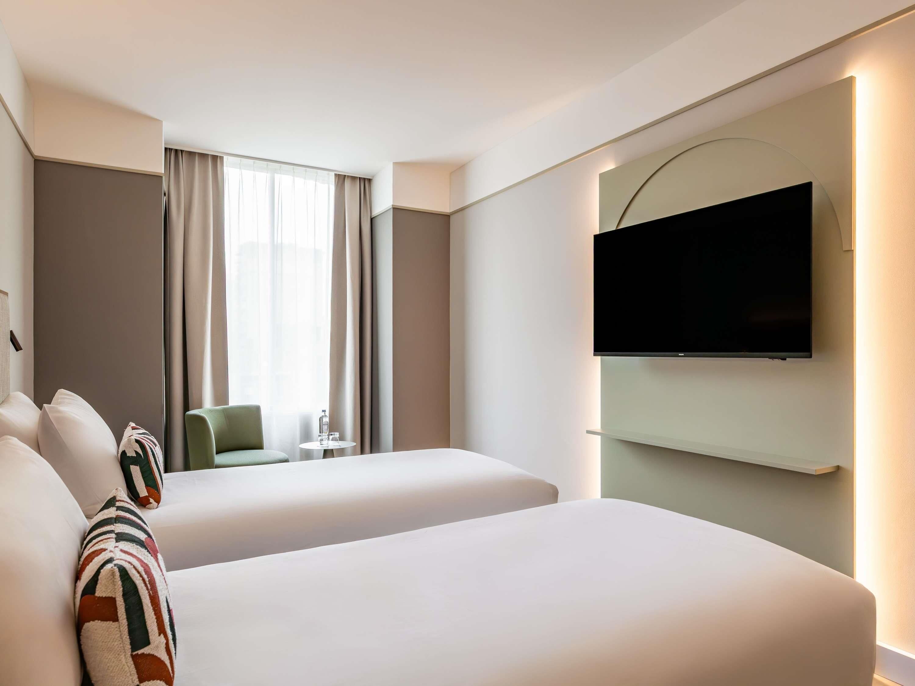 Ein Hotelzimmer mit zwei Betten, einem an der Wand montierten Fernseher, einem kleinen Tisch mit einem Stuhl und einem Fenster mit Vorhängen.