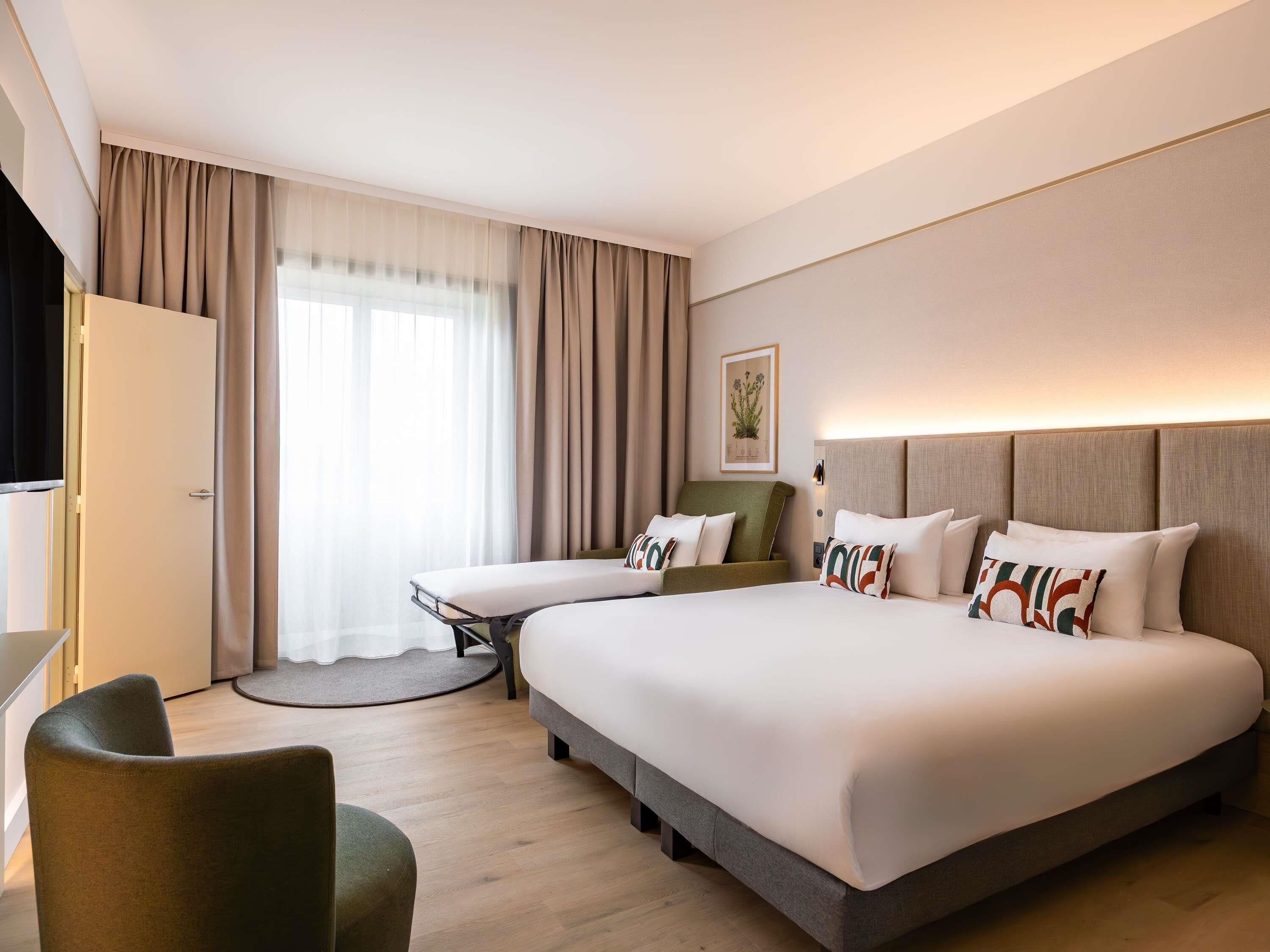 A hotel room with a large bed, a desk, a chair, and a window with curtains.