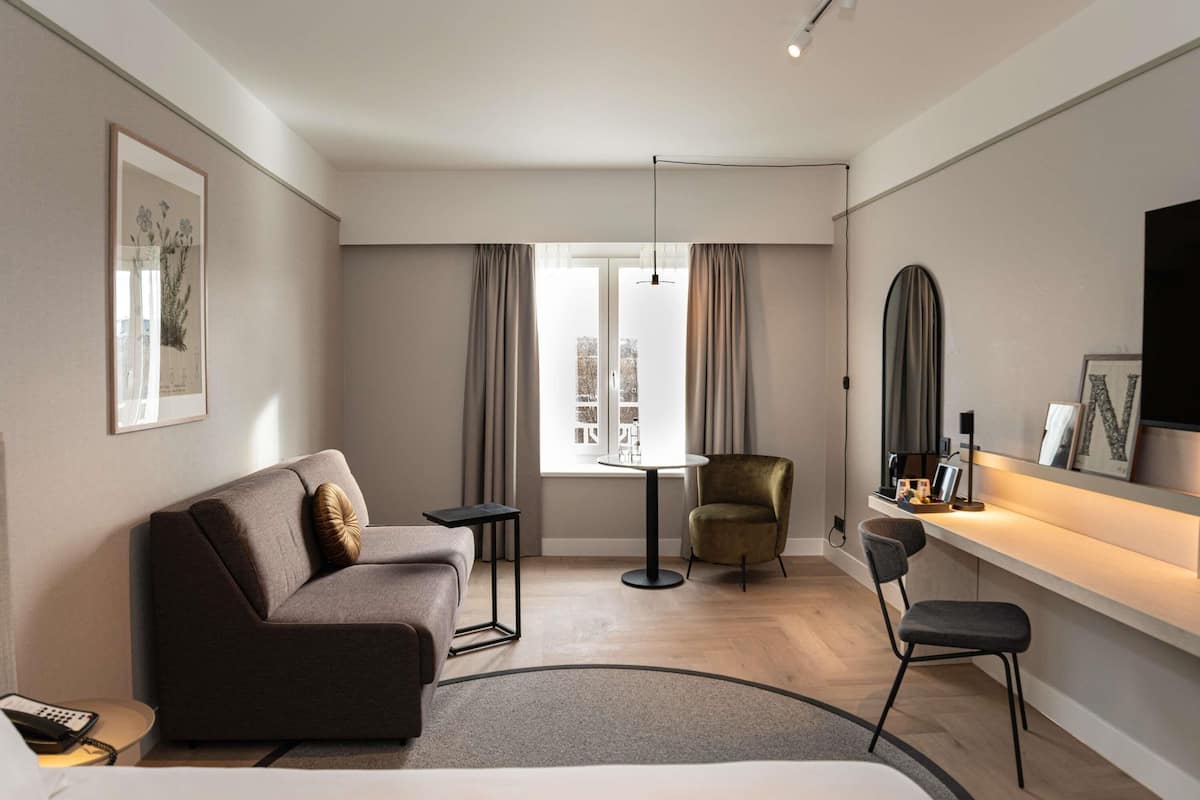 A modern hotel room with a sofa, a chair, a small table, a bed, a TV, and a window with curtains.