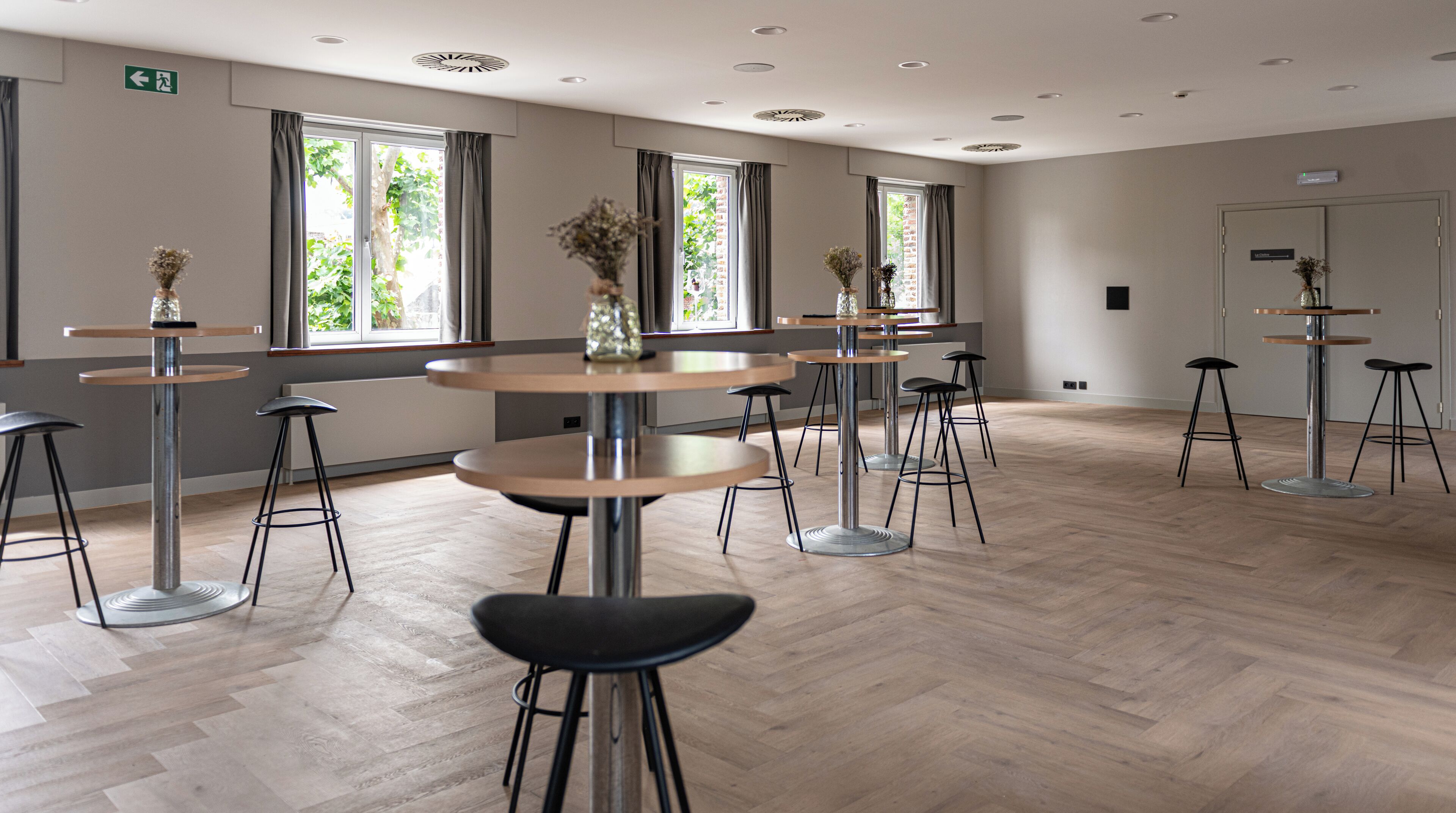 A spacious room with multiple round tables and black stools, large windows with curtains, and a herringbone-patterned wooden floor.