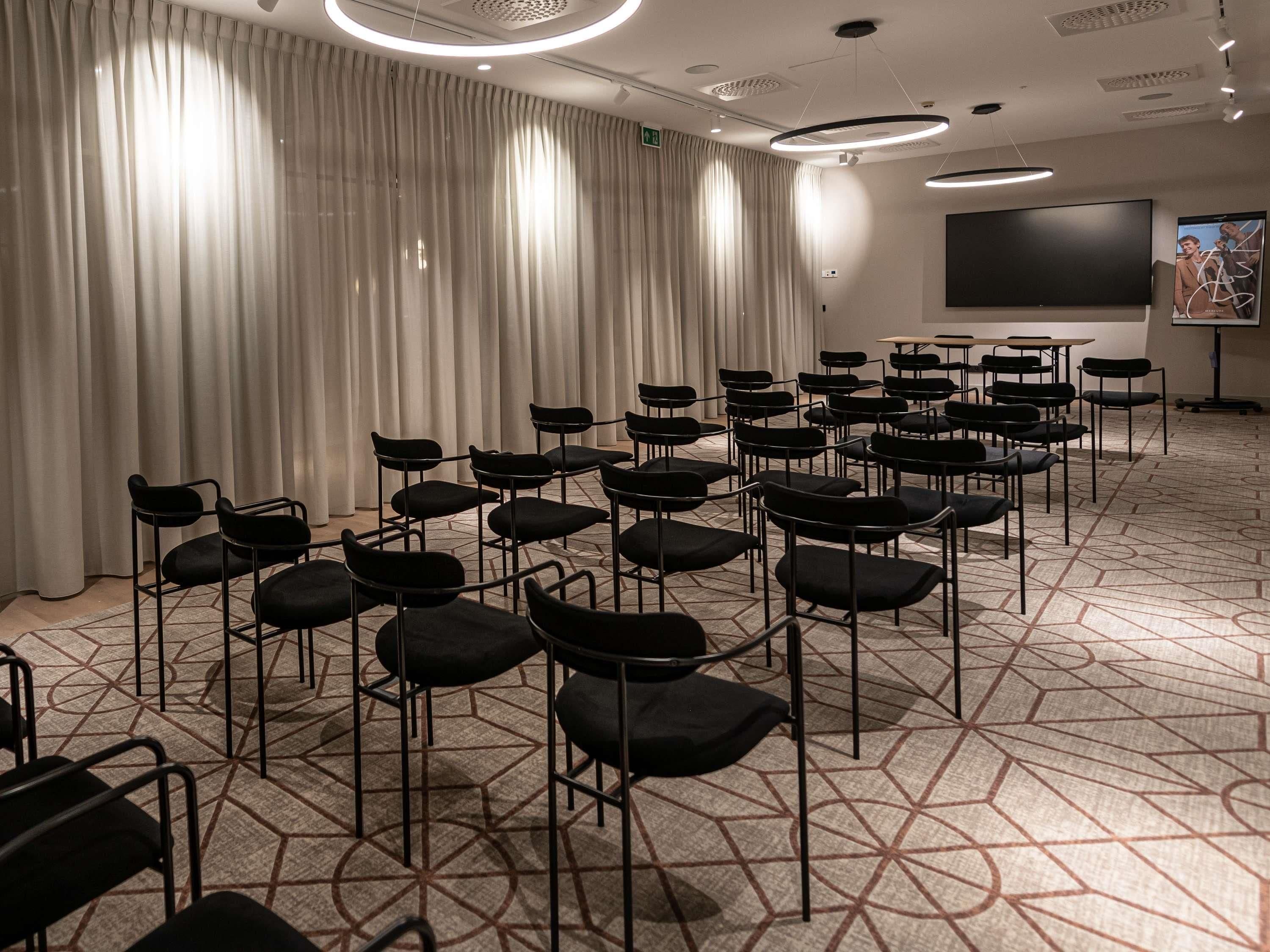 Een vergaderzaal met rijen stoelen, een groot scherm en een kamerbreed tapijt met een patroon.
