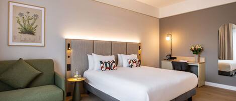 Habitación de hotel con cama, un sillón verde, un escritorio con lámpara, un espejo e ilustración botánica enmarcada en la pared.