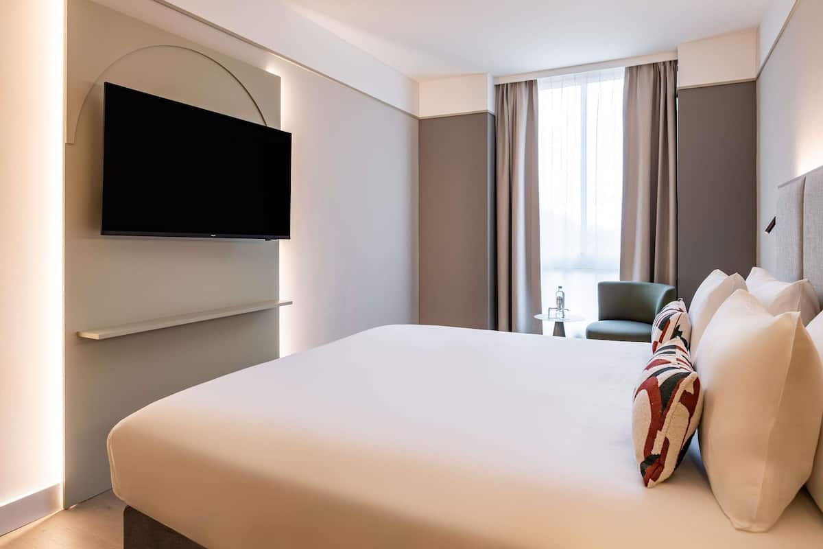 Una habitación de hotel moderna con una cama grande, un televisor de pantalla plana en la pared, una mesita pequeña y una silla.